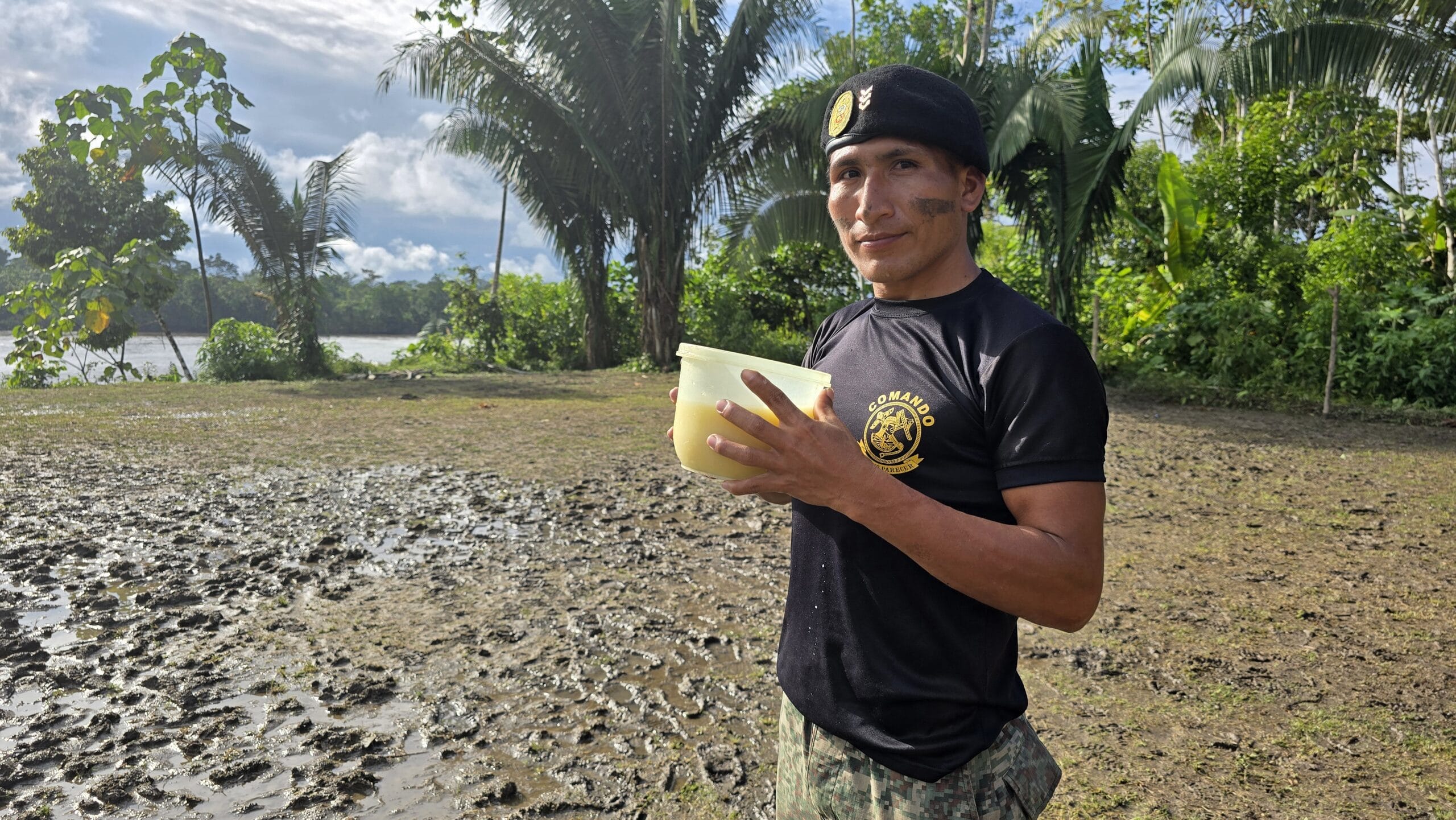 A man in a black shirt and hat stands outdoors near trees and muddy ground, holding a translucent container with a light-colored liquid.