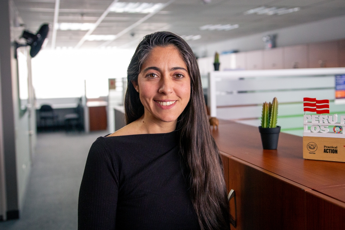 A woman with long dark hair and a black top stands smiling in a modern office near a wooden counter with a small cactus and a Peruvian flag decoration.
