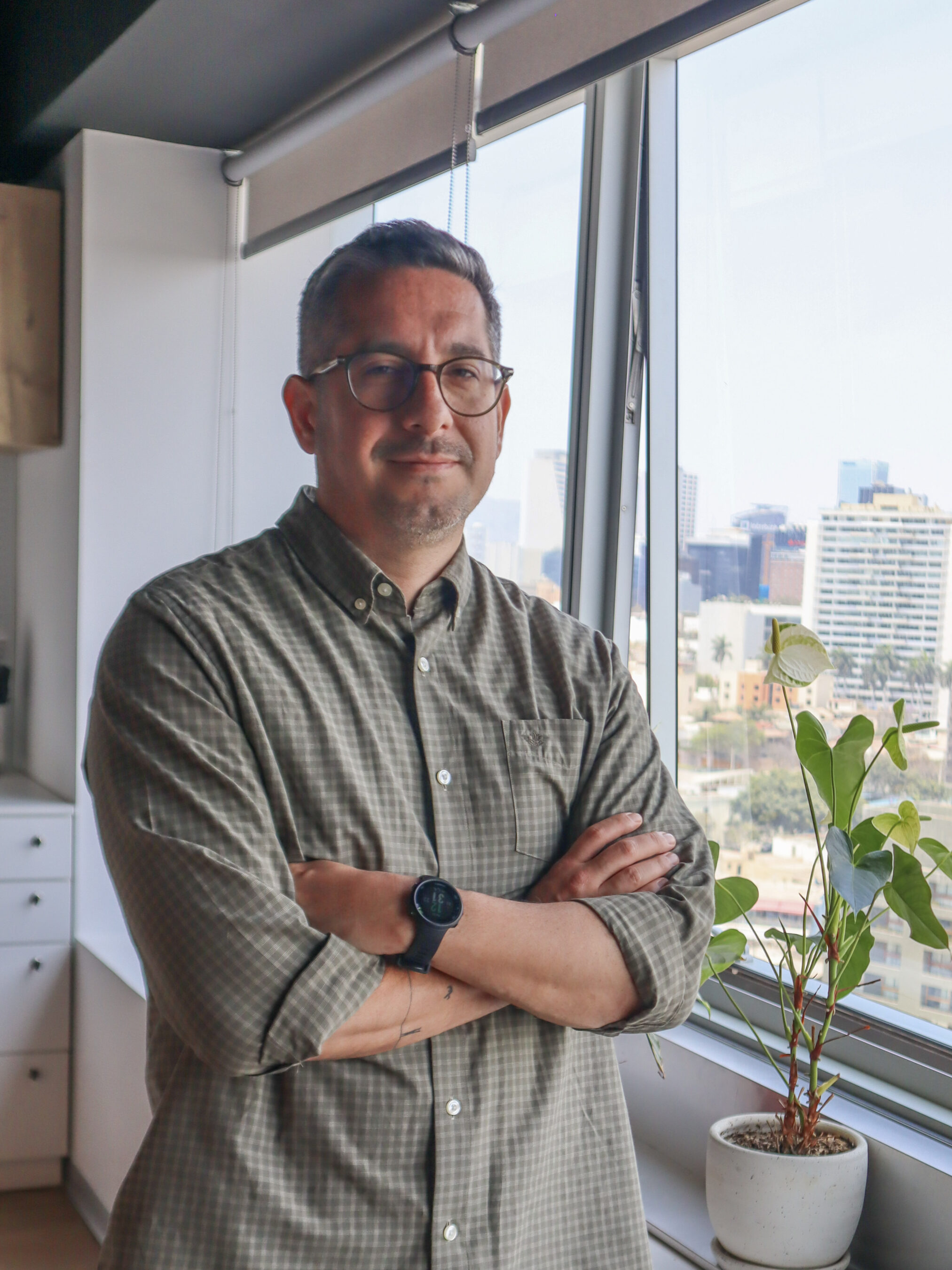 A man with glasses and a smartwatch stands with arms crossed by a window in a modern office, with city buildings visible outside and a potted plant on the windowsill.
