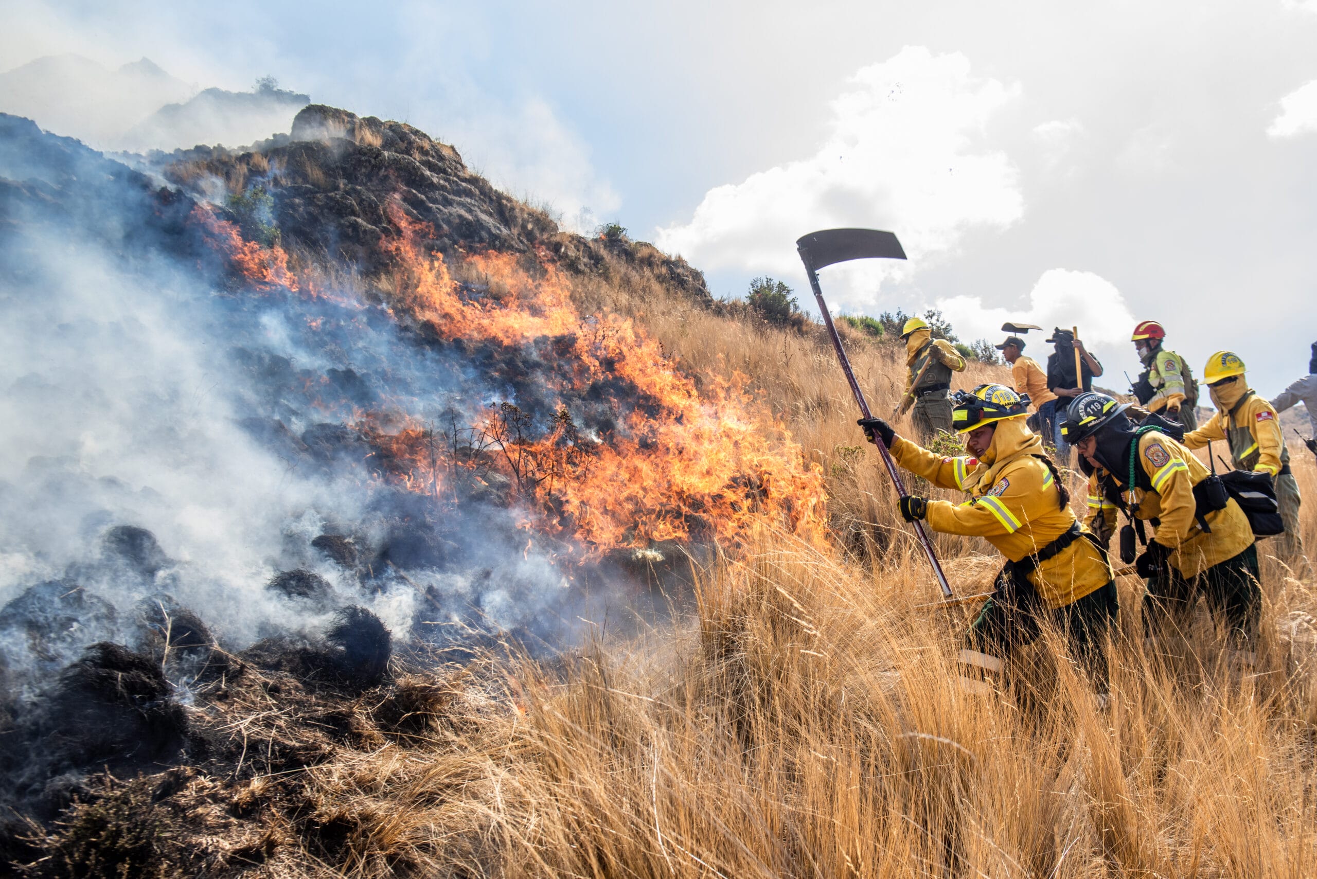 Firefighters in yellow protective gear work to contain a brush fire on a grassy hillside, using hand tools as flames and smoke rise in the background.