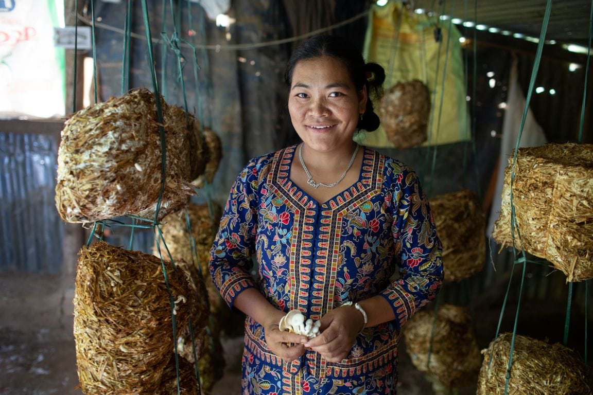 A woman stands smiling in a mushroom cultivation area, holding harvested mushrooms, with hanging mushroom substrates visible around her.