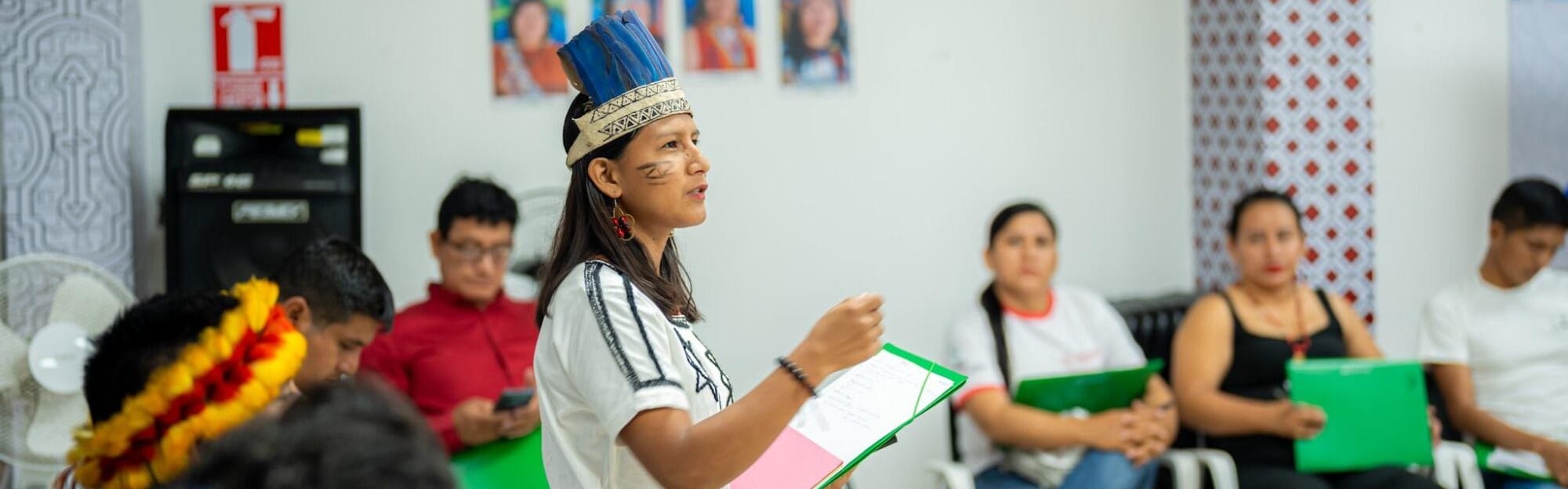 A woman wearing a traditional headdress speaks to a seated group, each holding green folders, in a brightly lit room.