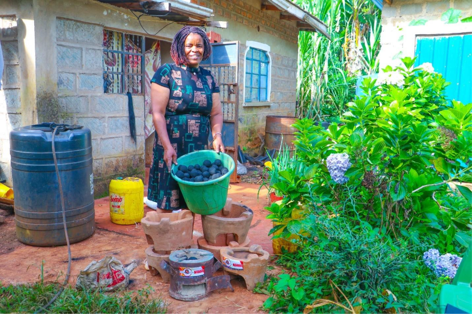 A woman stands outdoors holding a green bucket of charcoal balls next to two clay stoves, surrounded by plants and water containers near a brick house.