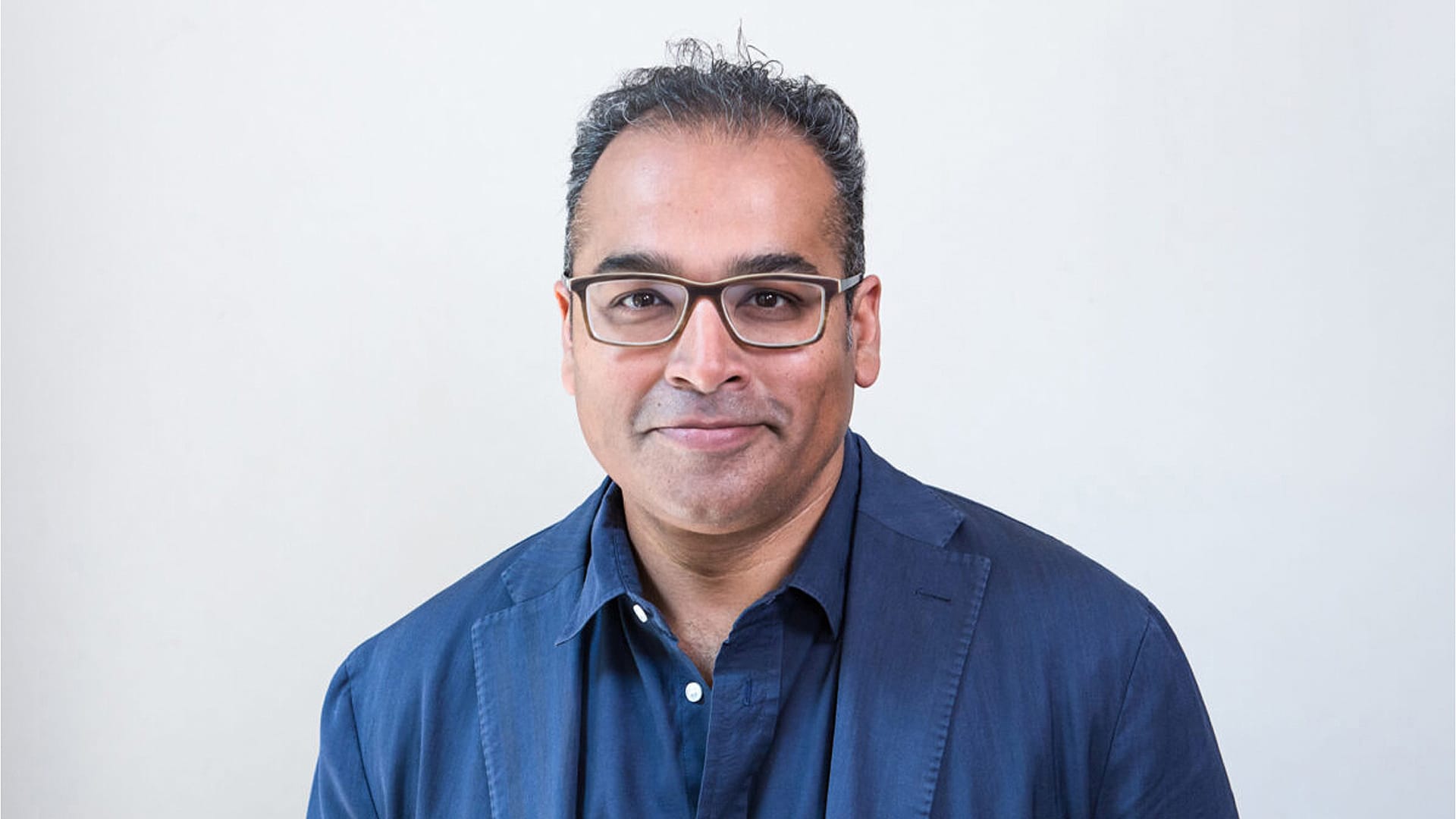 A man with short dark hair and glasses, wearing a blue blazer and a dark shirt, stands in front of a plain light-colored background.
