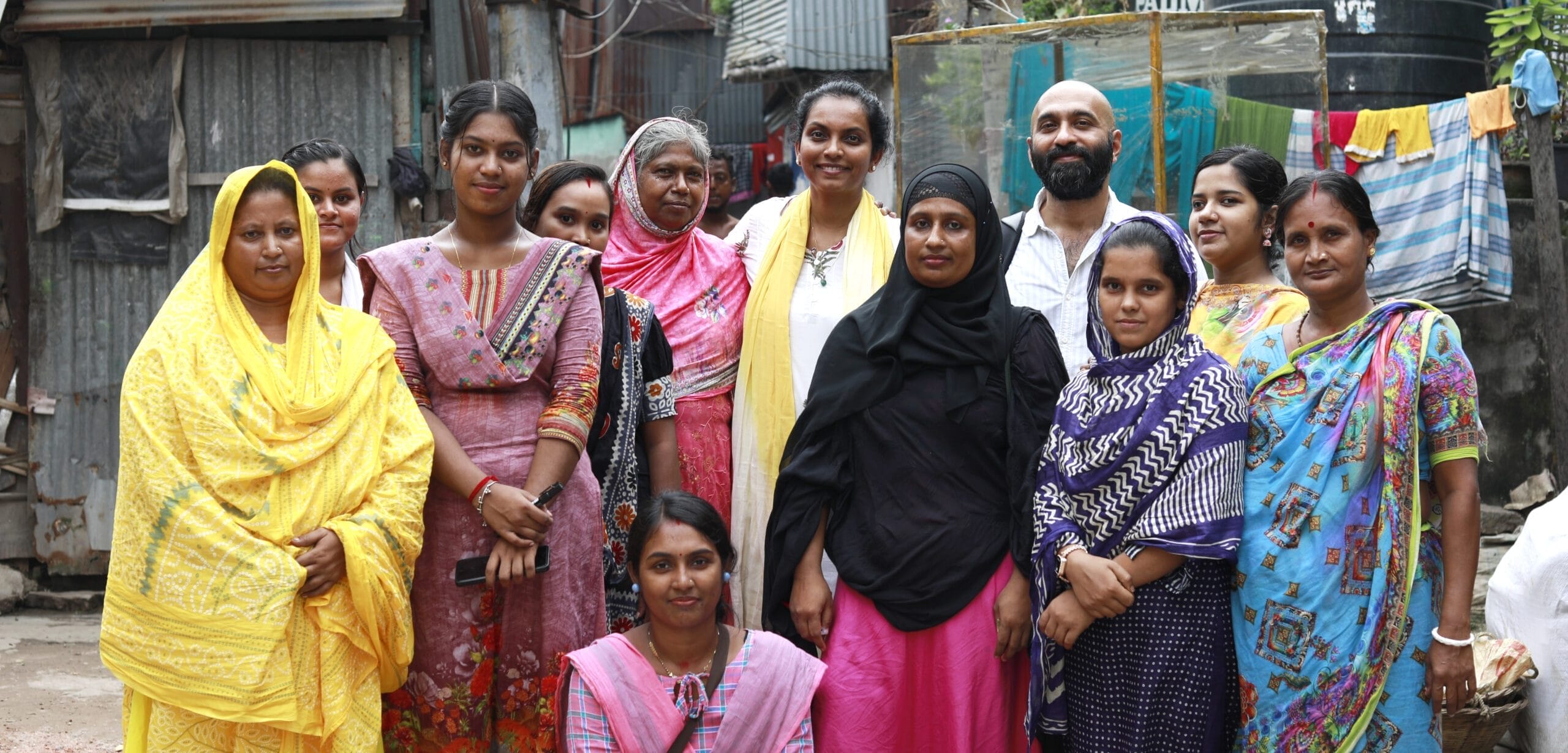 A group of thirteen adults, mostly women in colorful clothing, pose for a photo outdoors in an urban setting with laundry and corrugated metal structures in the background.