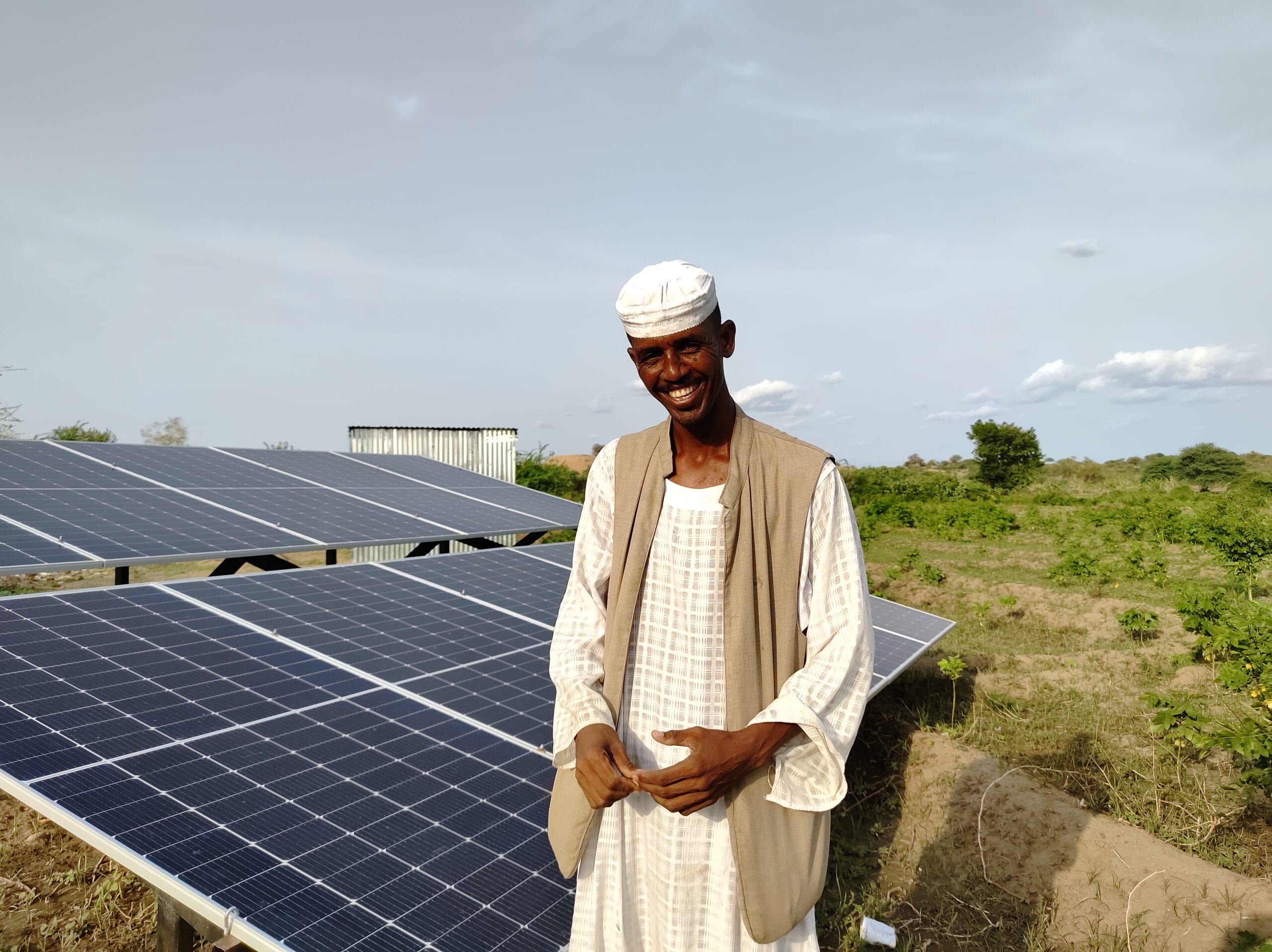 A man in traditional clothing stands outdoors next to a solar panel array, with green vegetation and a clear sky in the background.