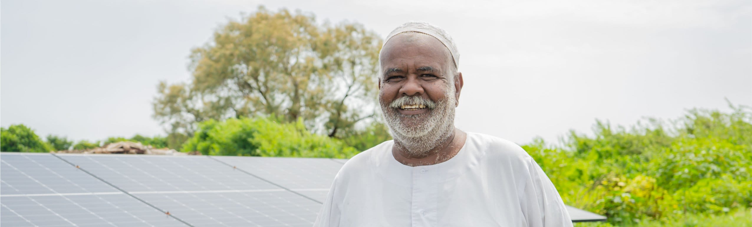 An older man in a white garment stands outdoors, smiling, in front of solar panels and greenery.