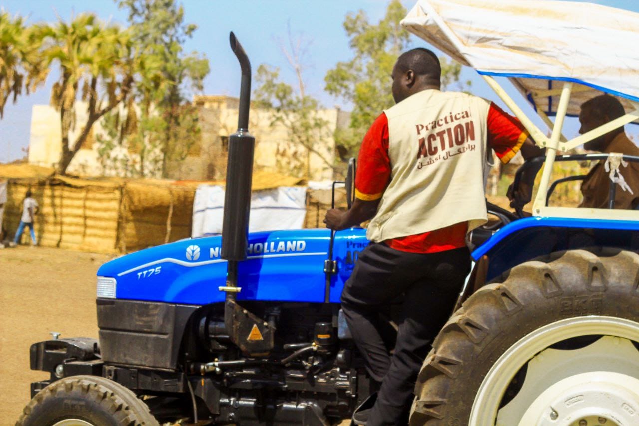 A man wearing a Practical Action vest sits on a blue New Holland tractor outdoors, with trees and buildings in the background.