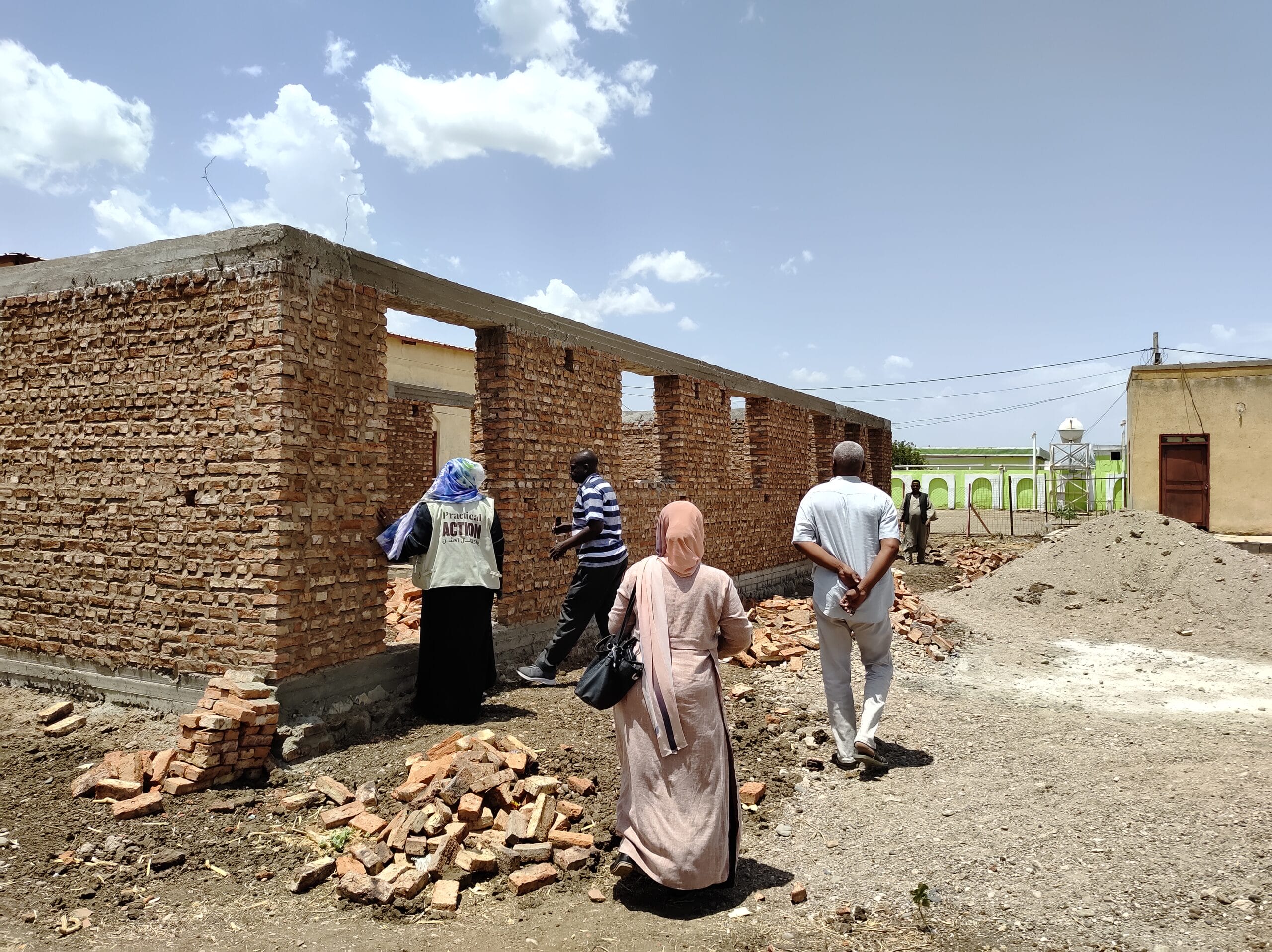 Four people walk around a partially constructed brick building under a bright sky, with scattered bricks and rubble on the ground.