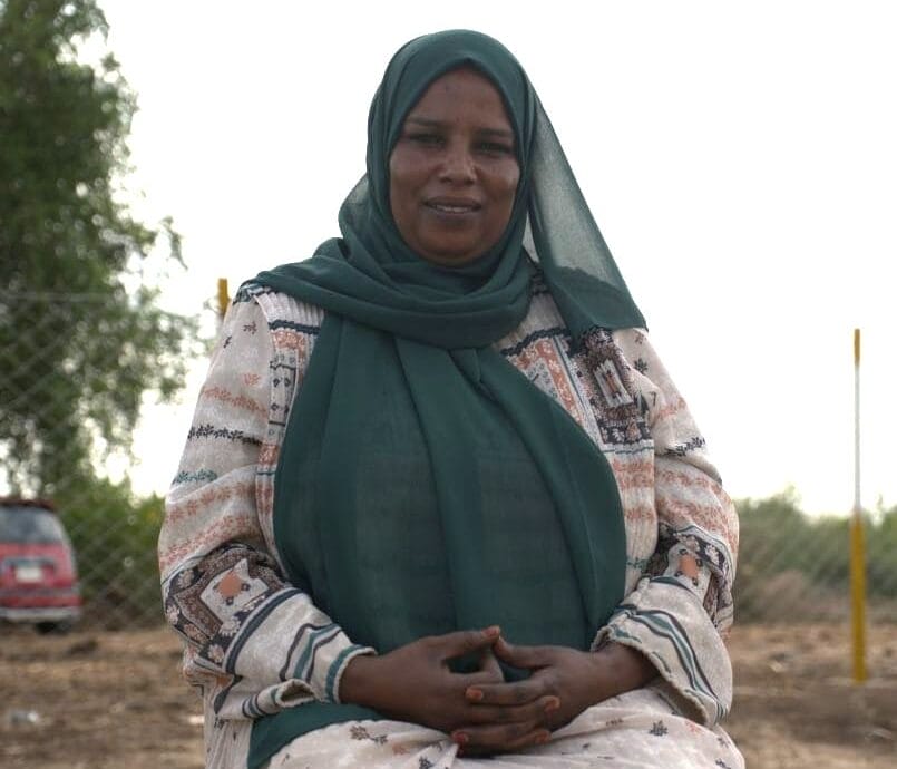 A woman wearing a patterned dress and green headscarf sits outdoors with her hands folded, a fence and trees in the background.