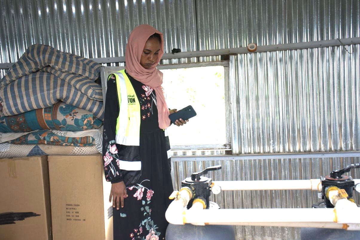 A woman in a safety vest and headscarf inspects equipment inside a metal-walled room with stacked mattresses and cardboard boxes.