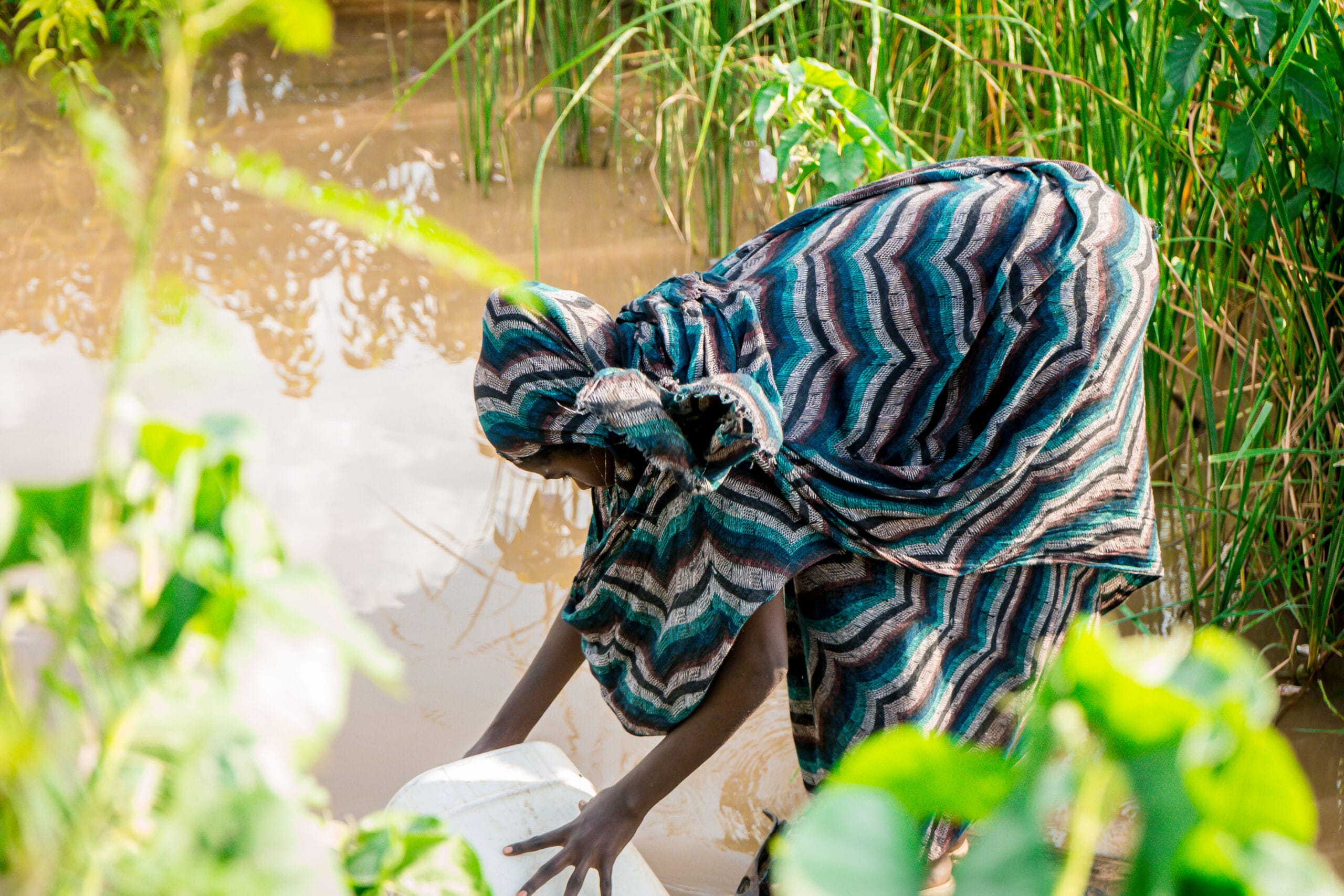A person in patterned clothing bends over near the edge of a muddy body of water, surrounded by green plants, while handling a white container.