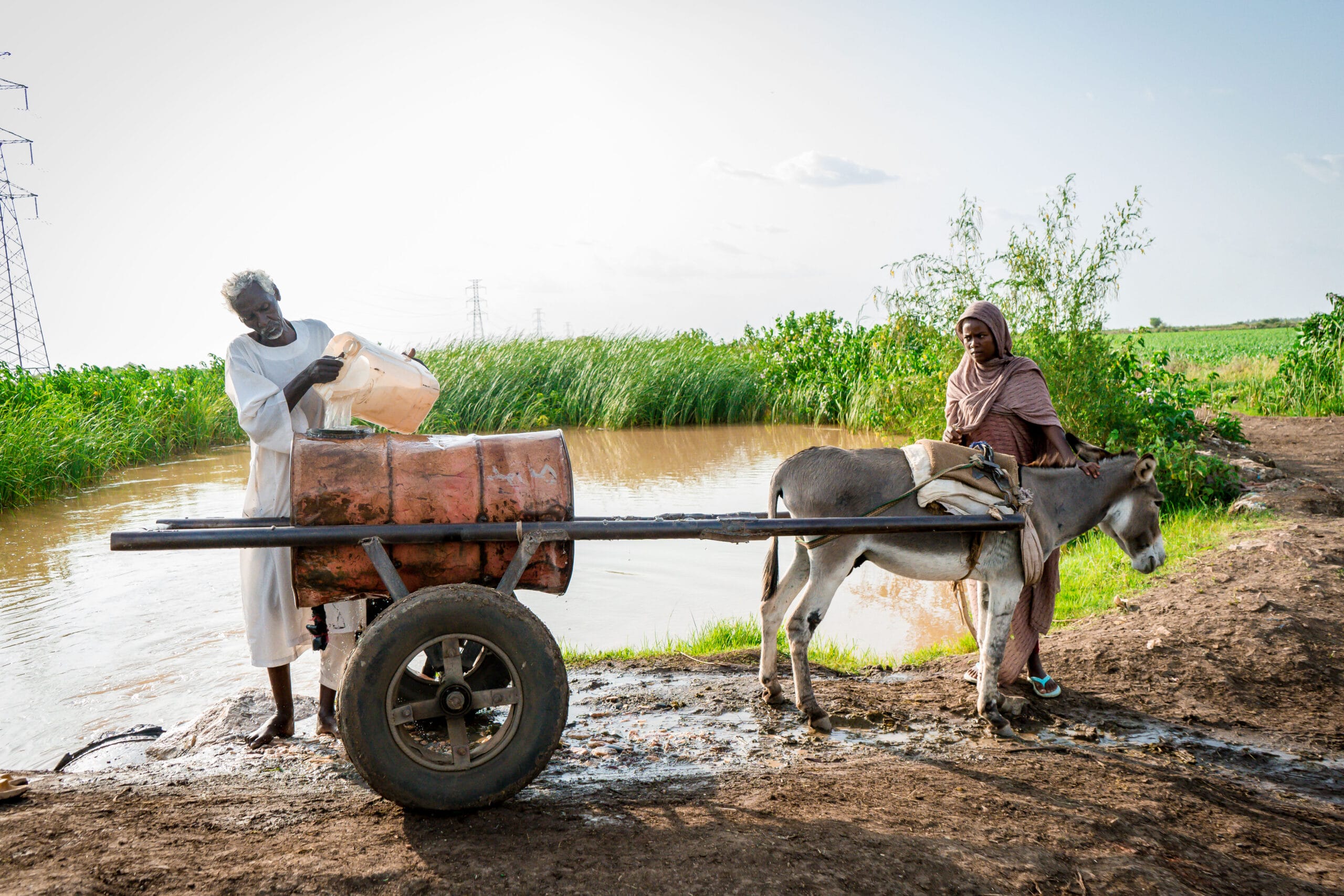 A man pours water from a container into a drum on a donkey cart beside a river, while a woman stands nearby holding the donkey's reins.