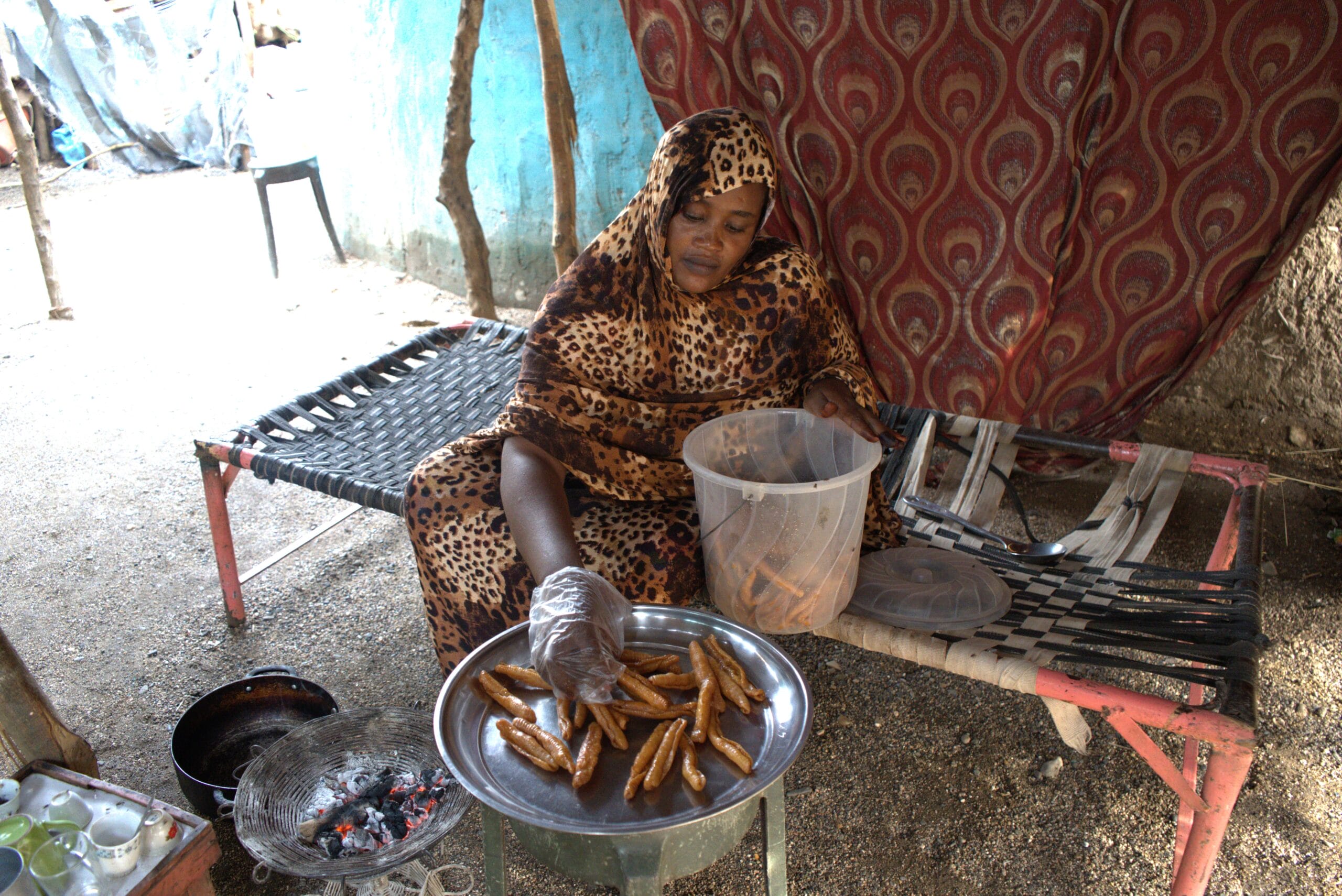 A woman in patterned clothing sits on a woven bed, placing fried pastries from a plastic container onto a metal tray. Cooking supplies and a charcoal grill are nearby.