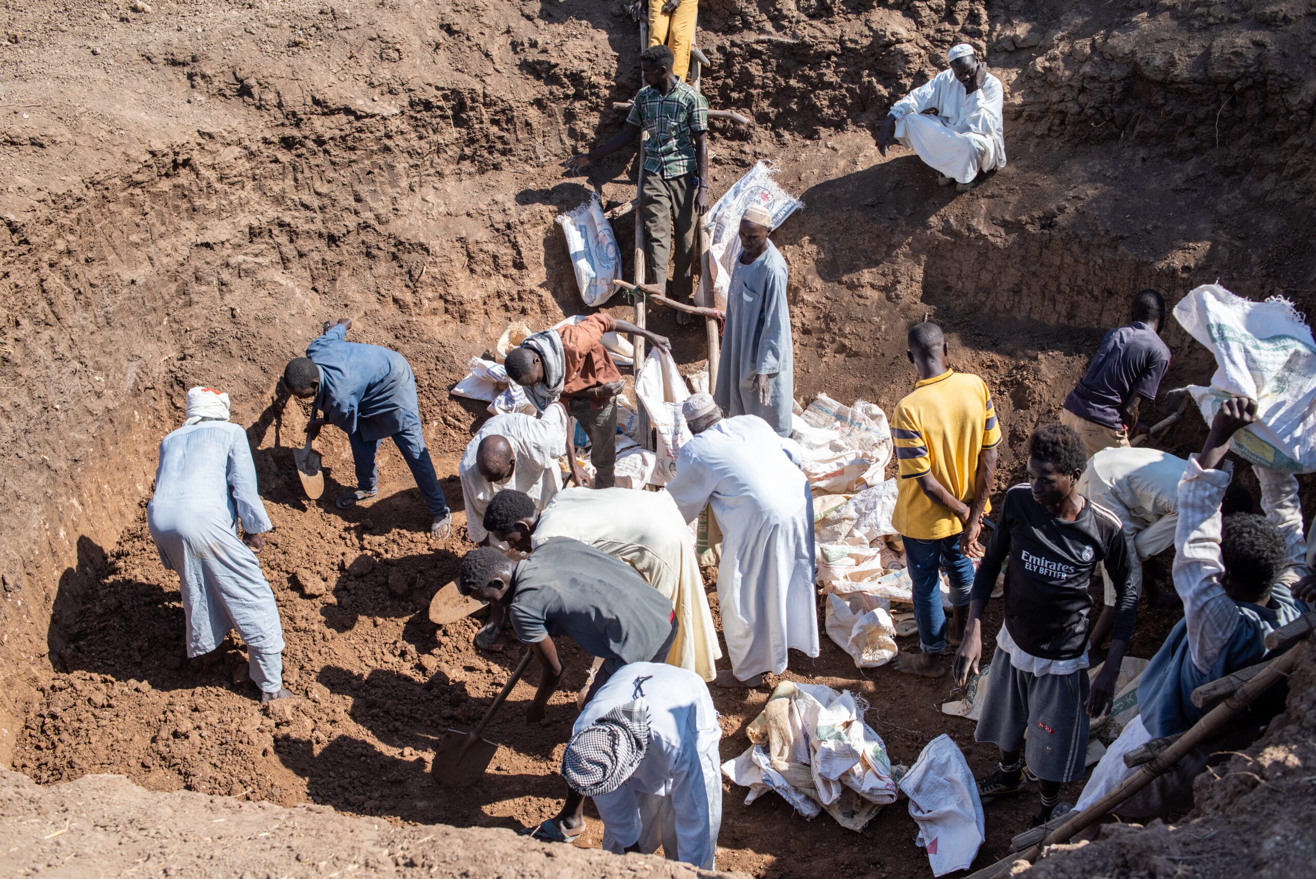 A group of people are gathered in a large dirt pit, some shoveling earth, while others stand around or place bags on the ground.