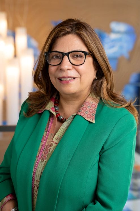 A woman with long brown hair and glasses, wearing a green blazer and patterned shirt, stands indoors with abstract blue artwork in the background.