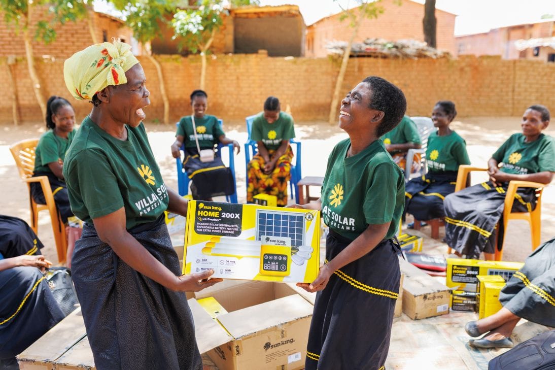 Two women in matching green shirts exchange a boxed solar home kit while others sit in a circle outdoors, smiling and wearing similar attire.