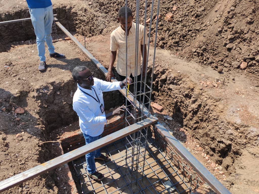 Two men stand in a construction site, inspecting vertical steel reinforcement bars set in a foundation trench.