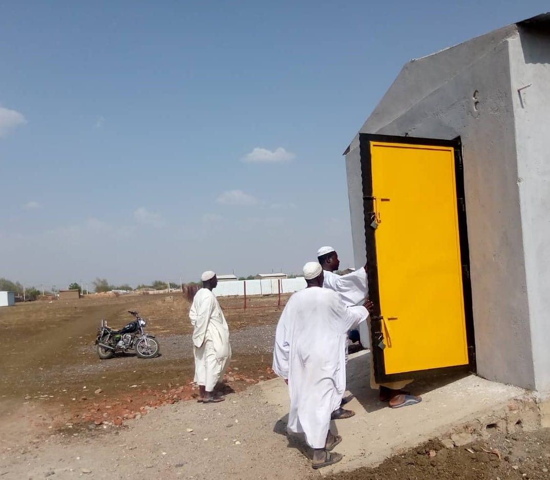 Three men in white robes stand near a small gray building with a bright yellow door; one man is entering while two others watch. A motorcycle is parked in the background.