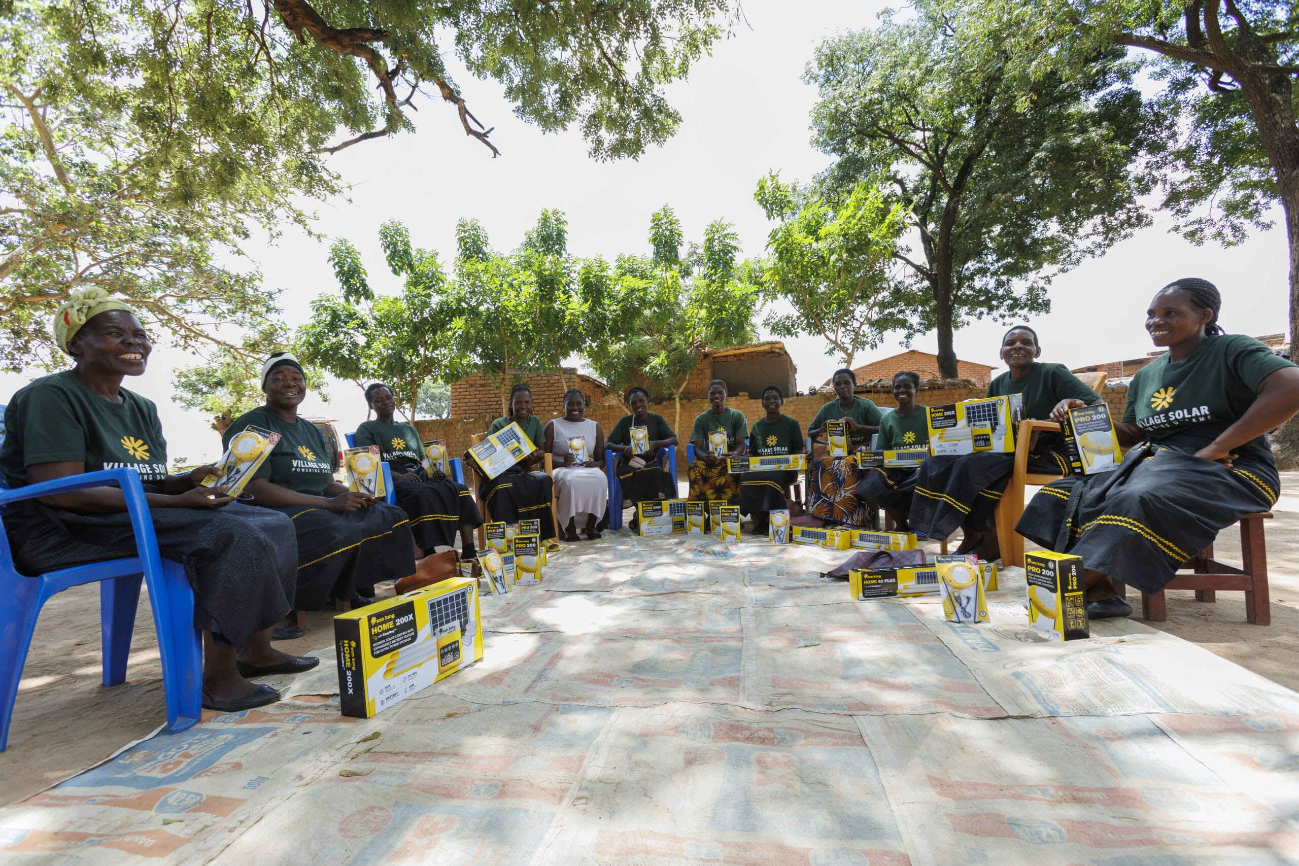 A group of women sit in a circle outdoors, each holding a box and booklet, with similar boxes placed on the ground around them. Trees and buildings are visible in the background.