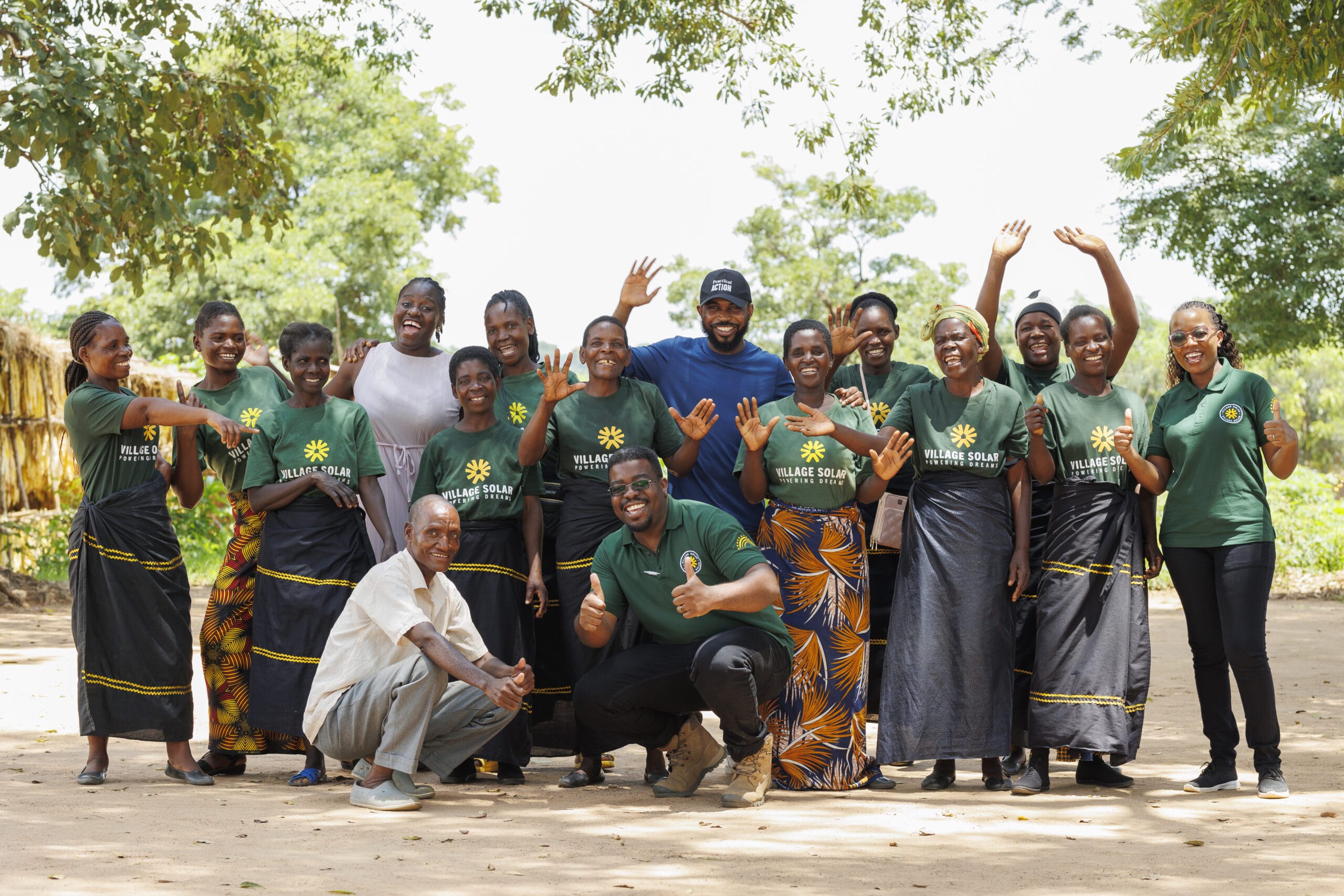A group of people pose and smile outdoors, with some standing and others kneeling, most wearing matching green "Village Solar" shirts. Trees and greenery appear in the background.