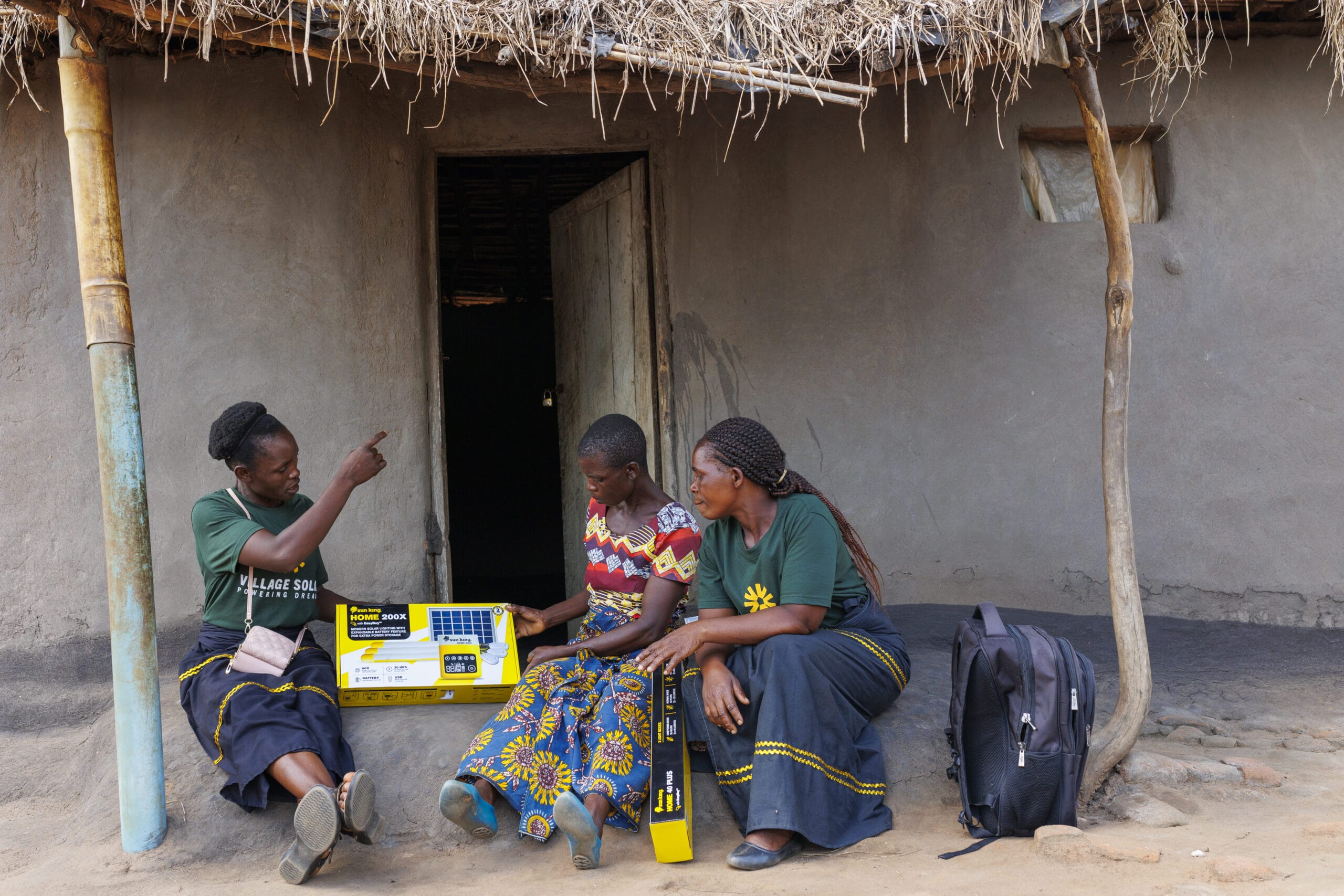 Three women sit outside a mud house; one points while another holds a solar home system kit box, and the third listens, with a backpack on the ground nearby.