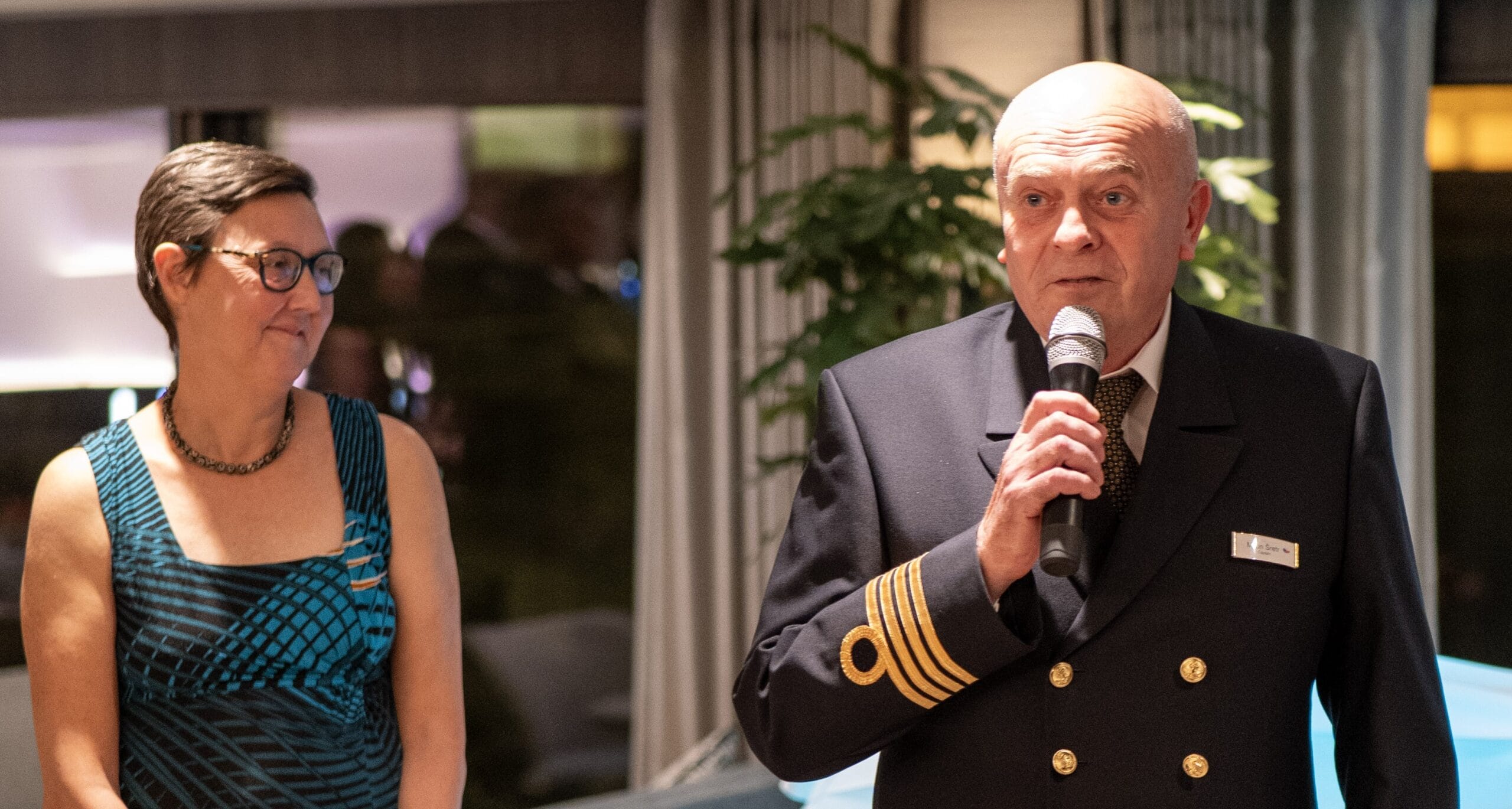 A man in a captain's uniform speaks into a microphone while a woman in a blue patterned dress stands beside him, both indoors.