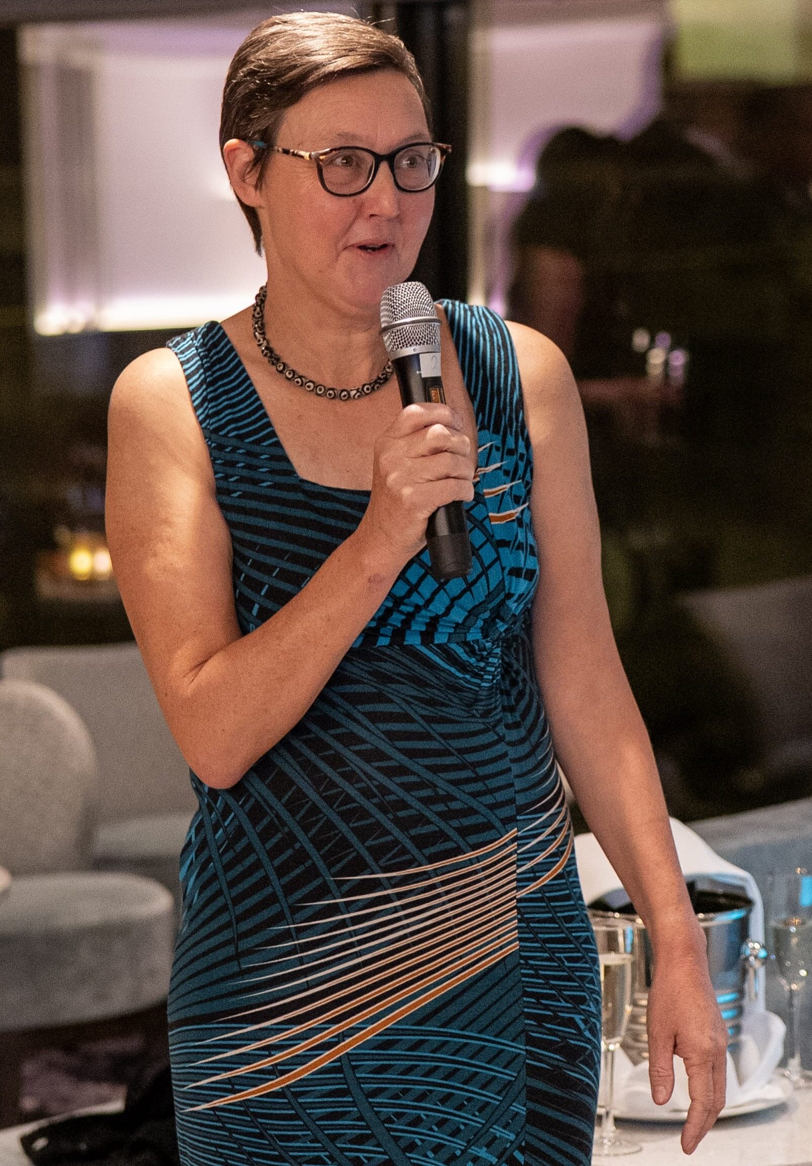 A woman in a patterned dress speaks into a microphone at an indoor event, with tables and glasses visible in the background.