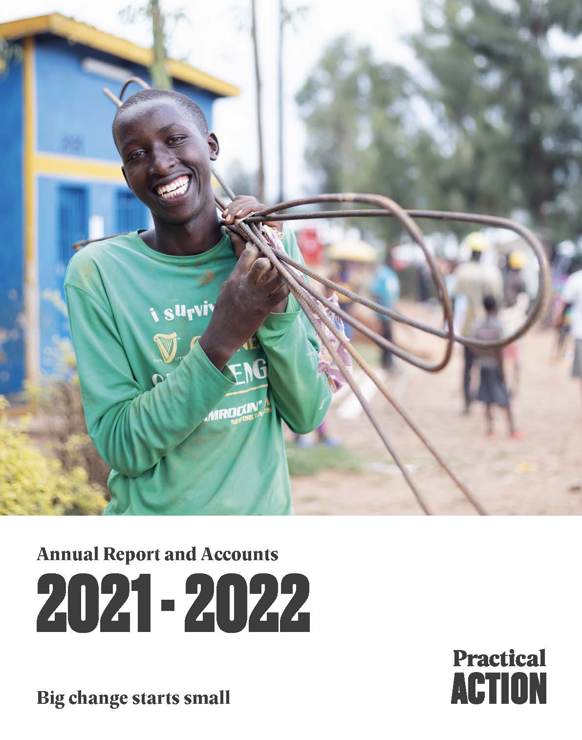 A smiling young person in a green shirt carries metal rods on their shoulder in an outdoor setting, with buildings and other people visible in the background.