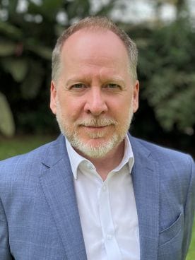 A middle-aged man with a short beard and light hair, wearing a blue blazer and white shirt, standing outdoors with greenery in the background.