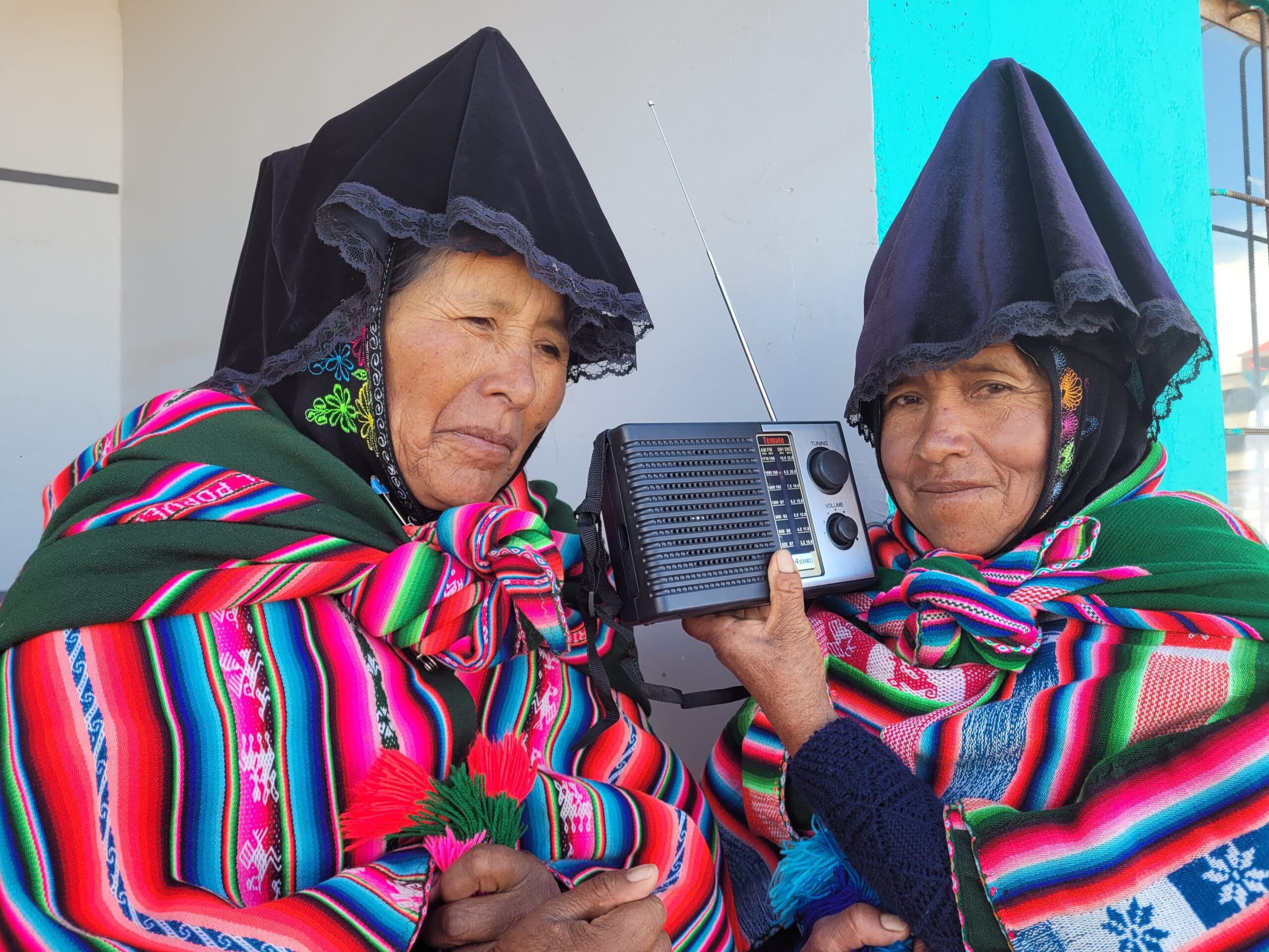 Two elderly women wearing traditional Andean clothing and head coverings hold a portable radio between them, standing against a brightly colored wall.