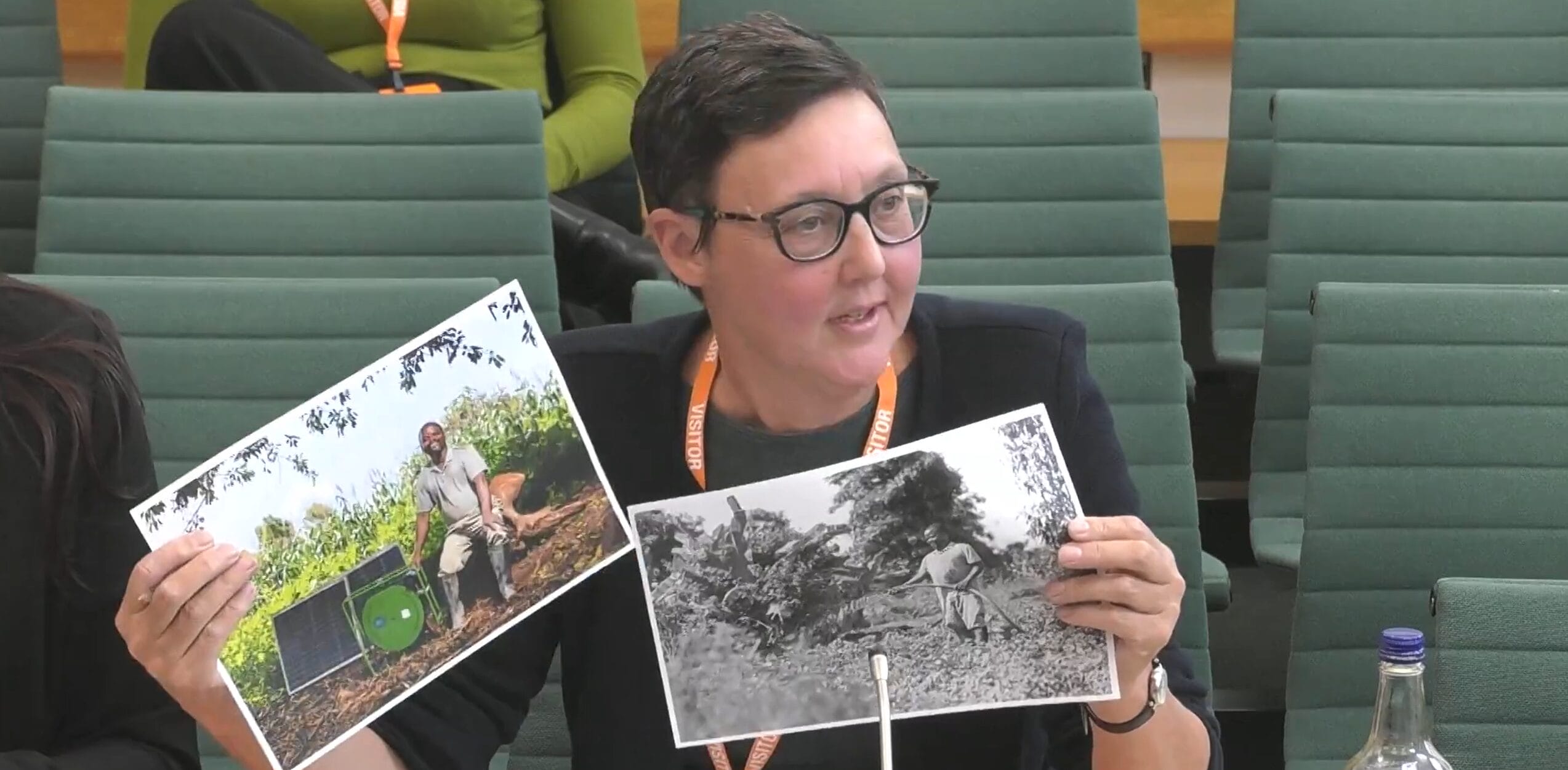 A person seated in a conference room holds up two photos: one in color showing a man outdoors with equipment, and one in black and white showing a person working in a field.