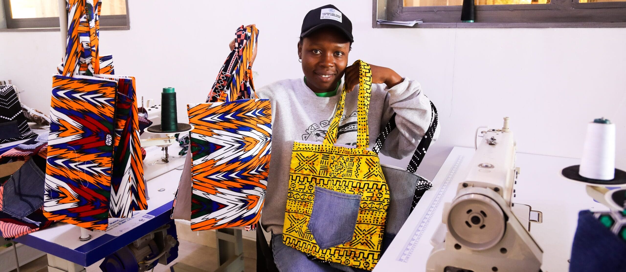 A person sits at a sewing machine in a workshop, displaying colorful patterned bags, with more bags and sewing materials on the table.