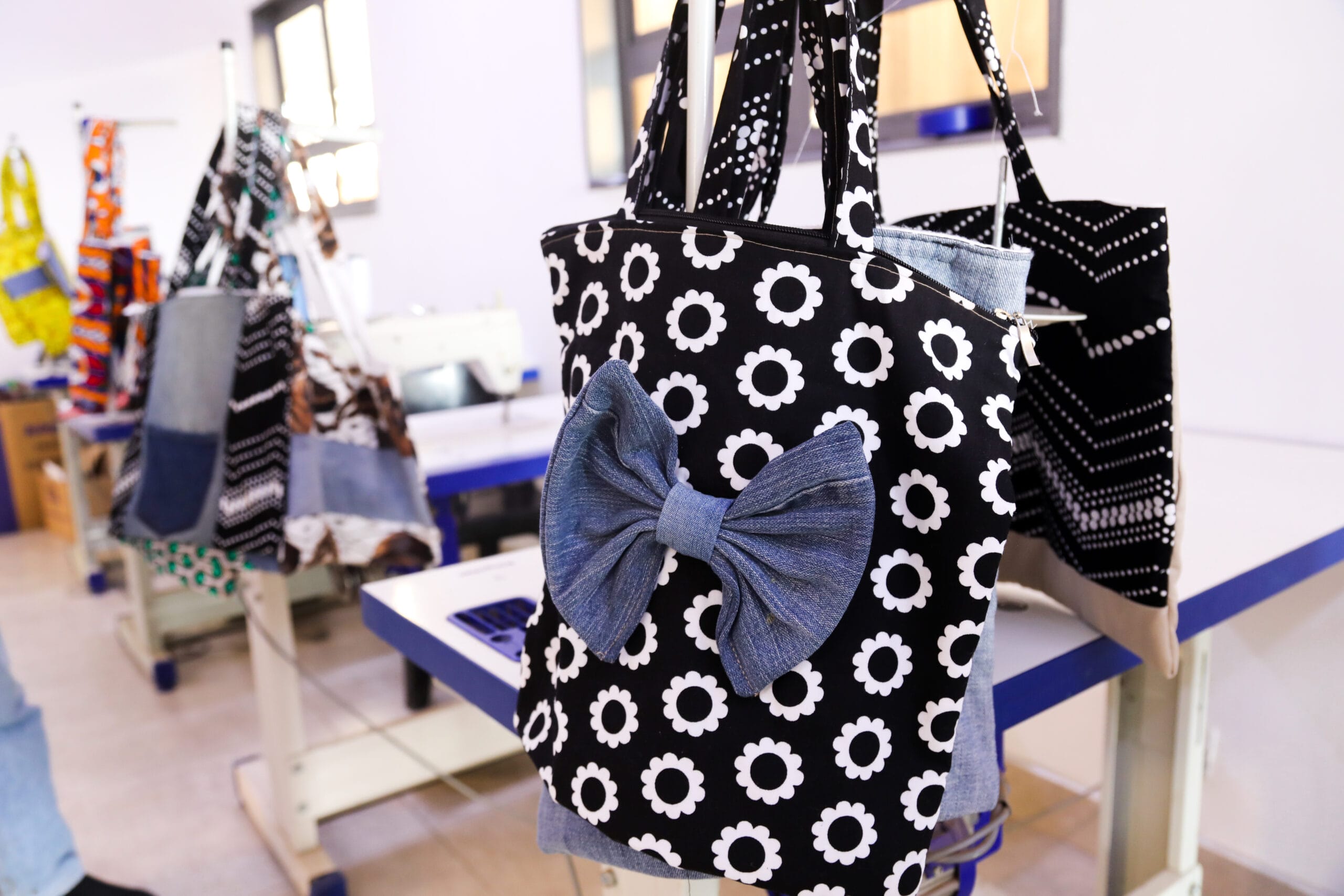 A black tote bag with white floral patterns and a large blue denim bow is displayed in a sewing or craft room.