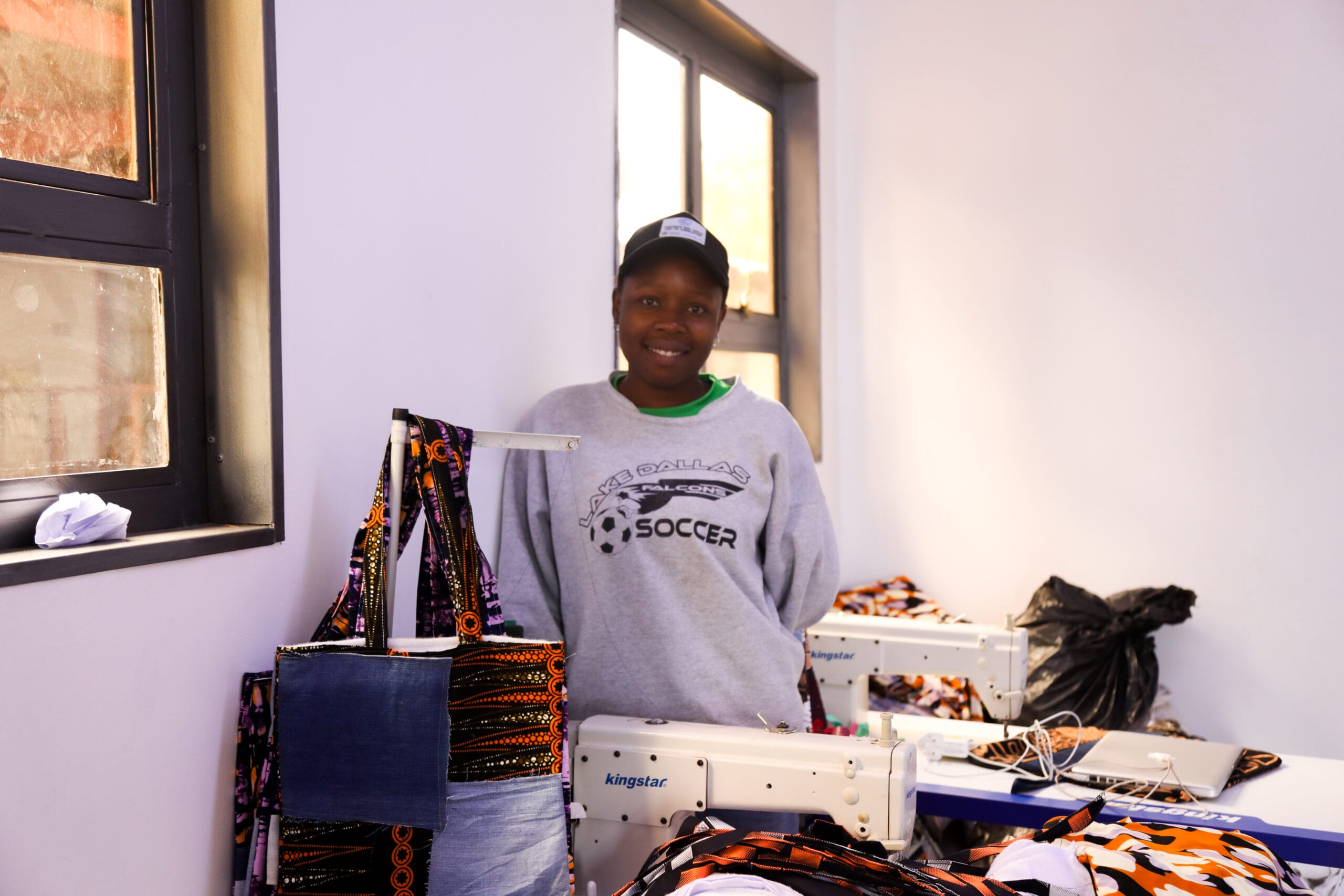 A person wearing a gray "Soccer" sweatshirt and cap stands in a room with sewing machines, colorful fabric, and finished bags.