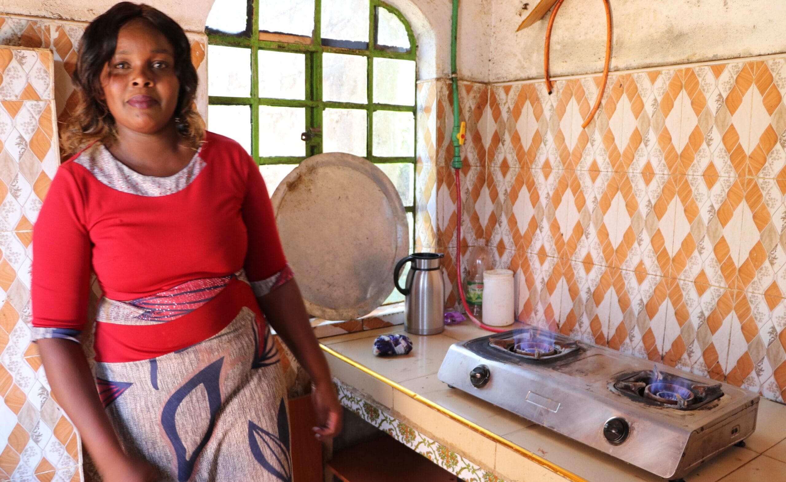 A woman stands in a kitchen next to a two-burner gas stove with blue flames, tiled walls, and various kitchen items on the counter.