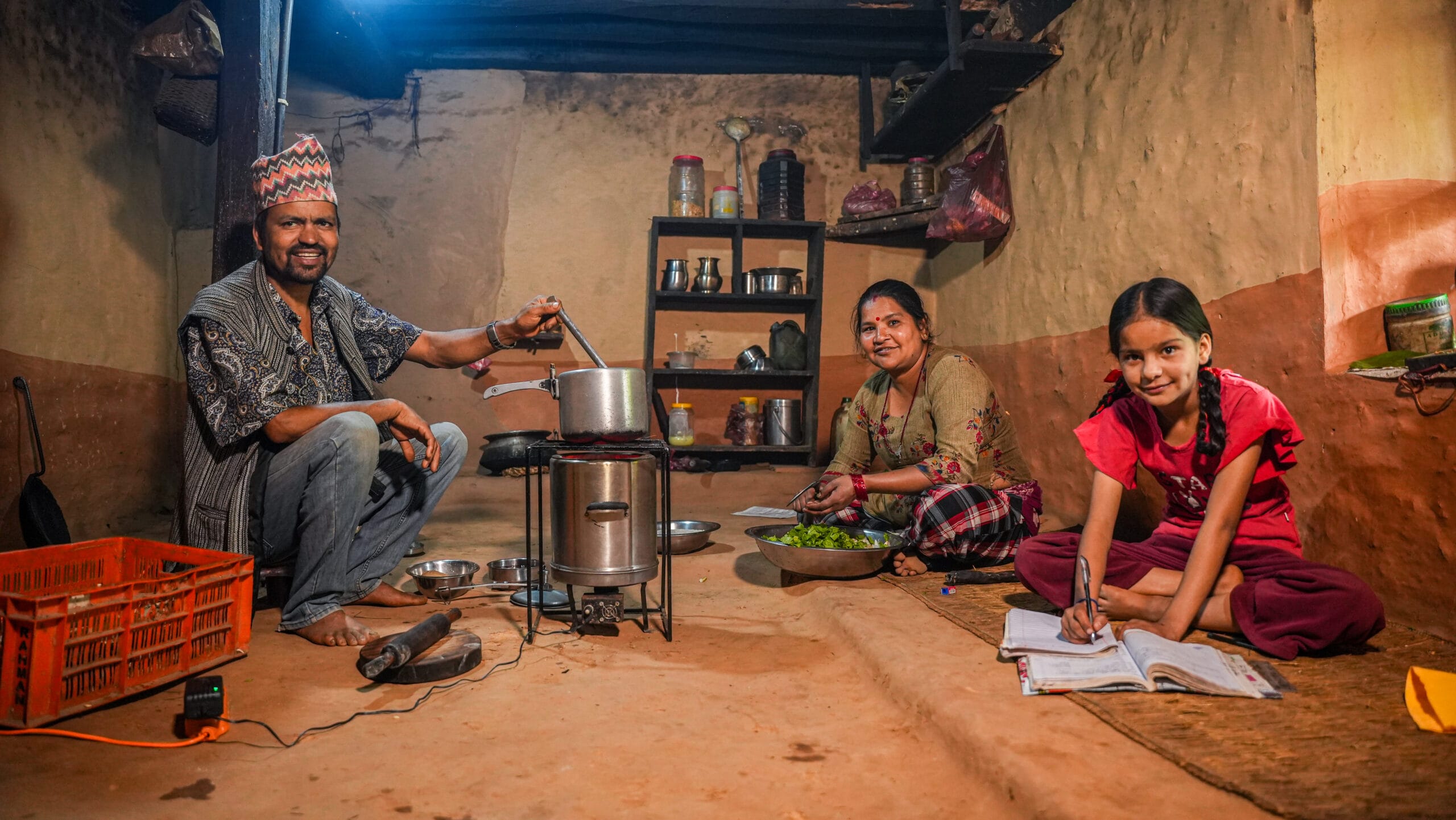 A man cooks on a stove, a woman prepares vegetables, and a girl does homework on the floor inside a modest kitchen with shelves and utensils.
