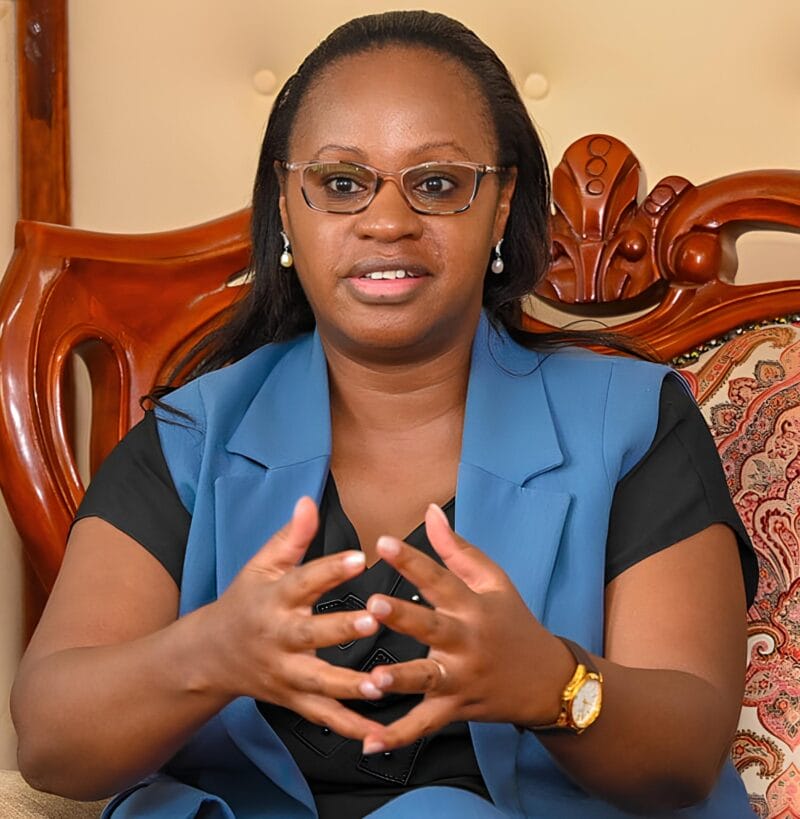 A woman wearing glasses and a blue vest sits on a carved wooden chair, gesturing with her hands while speaking.