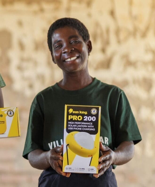 Three women stand indoors, smiling, and hold solar-powered product boxes while wearing matching "Village Solar" t-shirts.