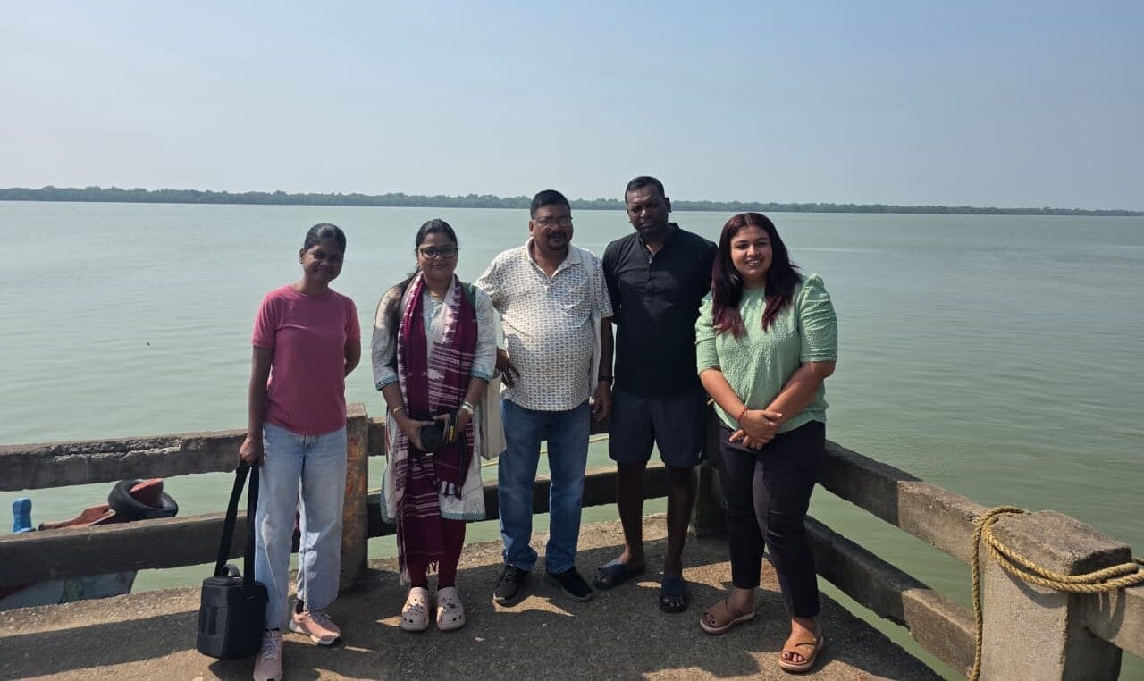 Five people stand on a concrete platform by a body of water under a clear sky, posing for a group photo.