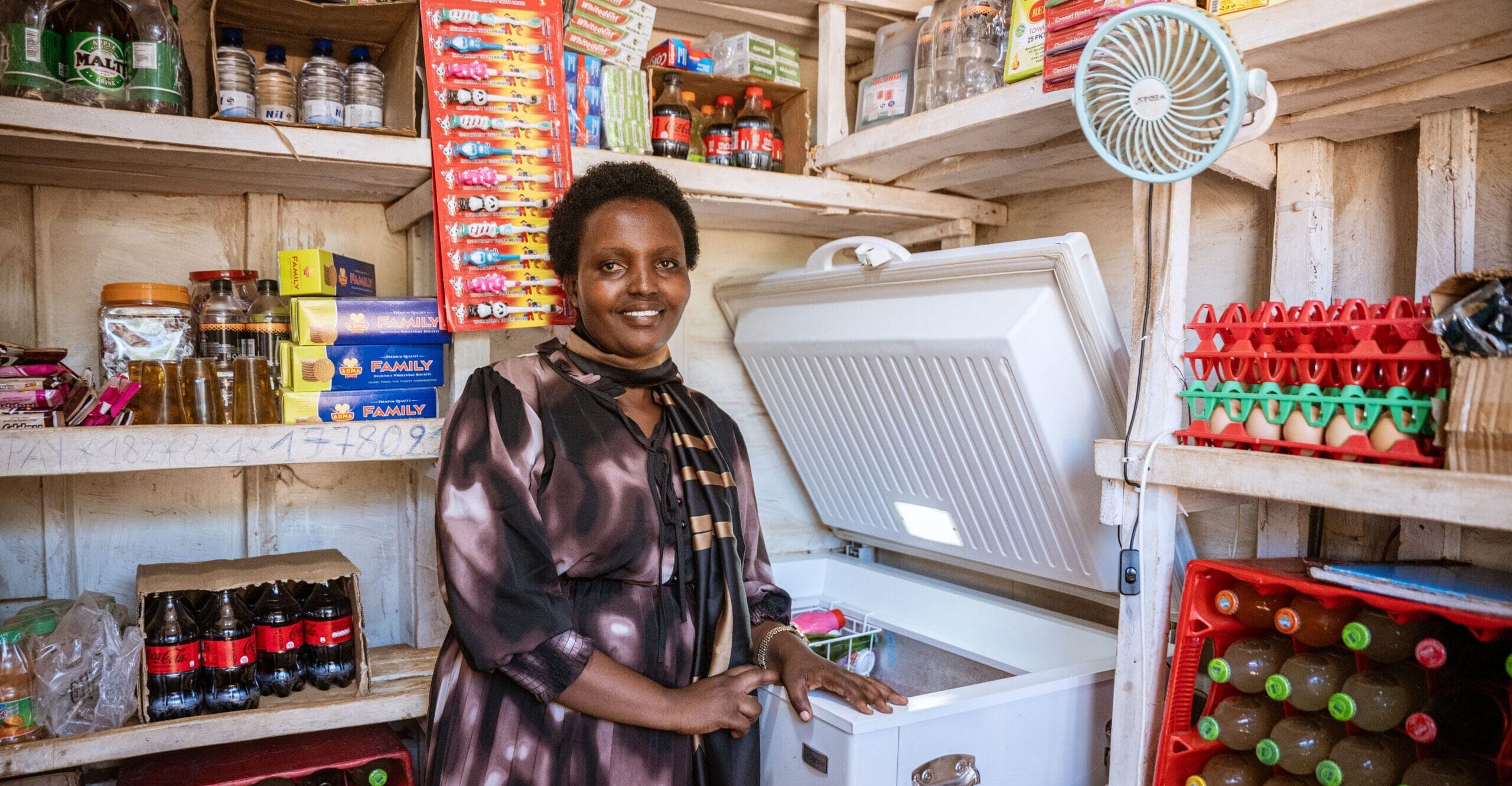 A woman stands smiling in a small shop, surrounded by shelves stocked with drinks, eggs, and groceries. She is holding open a white chest freezer filled with items. The shop is neatly organised with various goods.