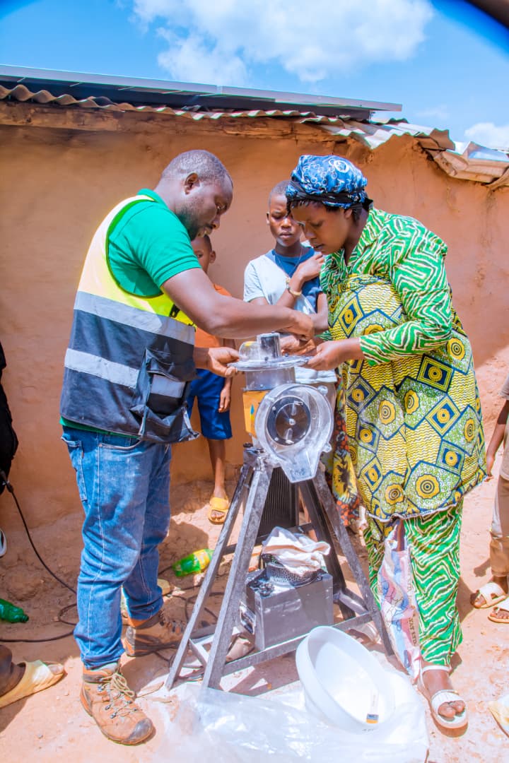 A man in a green shirt and safety vest helps a woman in a colorful patterned dress operate a food processing machine outdoors, with children and other people standing nearby.