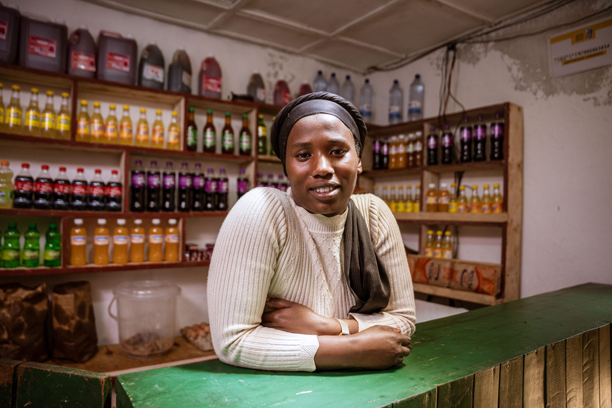 A woman wearing a cream jumper and headscarf leans on a green counter in a small shop, surrounded by shelves stocked with colourful drinks and bottled beverages.