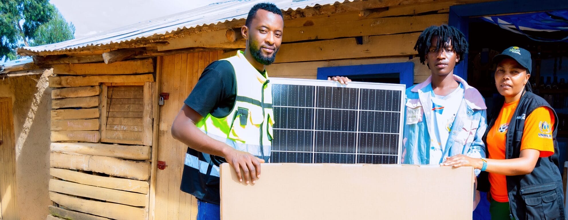 Three people stand outside a wooden building, holding a large solar panel together and looking at the camera.