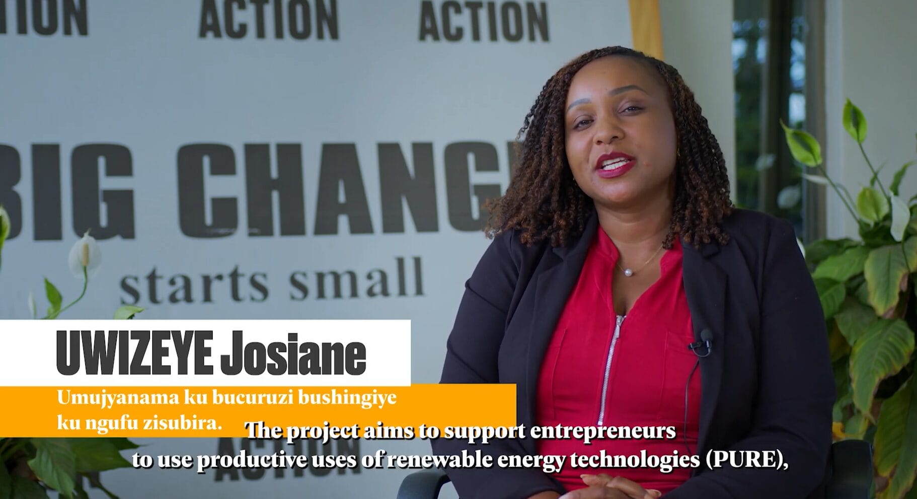 A woman in a red shirt and black blazer speaks in front of a wall with the words 