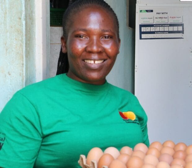A smiling woman in a green shirt holds a tray of eggs while standing in a doorway. Behind her, there is a large white egg incubator with eggs visible inside.