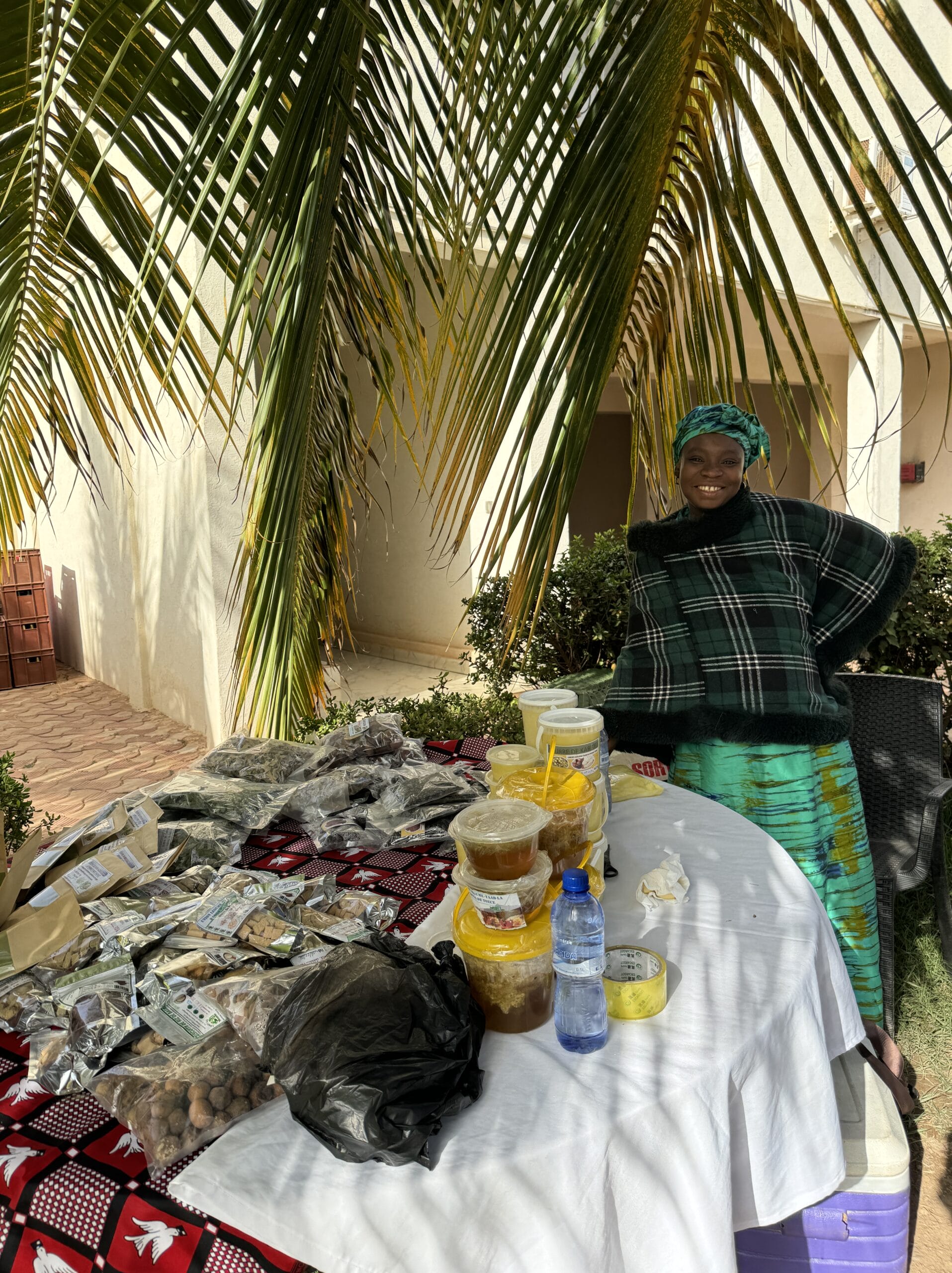 A woman wearing a green patterned outfit and headscarf stands smiling behind a table covered with jars, packets of food, and a water bottle under palm fronds, outside near a light-coloured building.