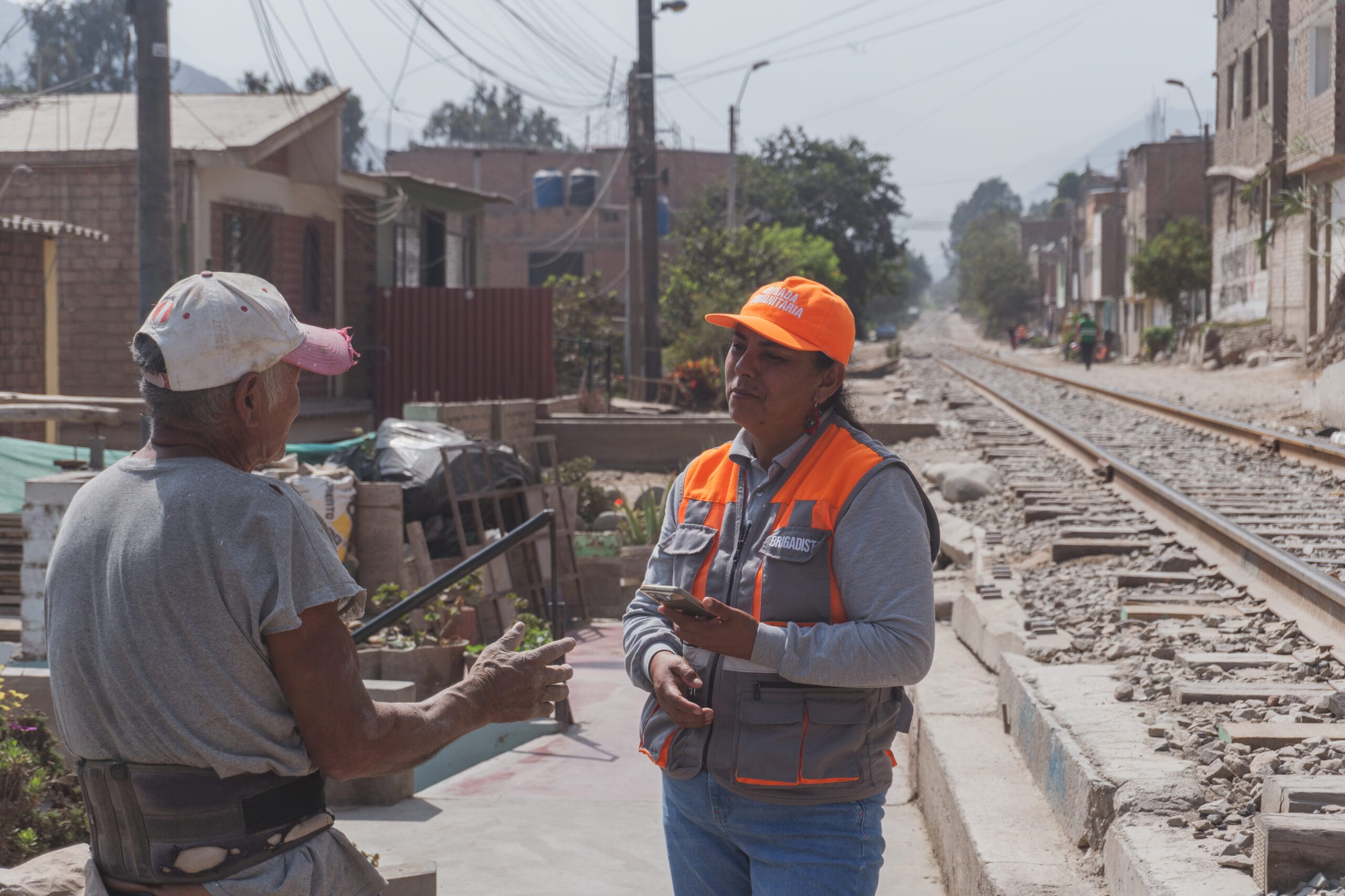 A woman in an orange safety vest and hat speaks with an older man near railway tracks under construction in a residential neighbourhood. She holds a clipboard while the man gestures with his hands.