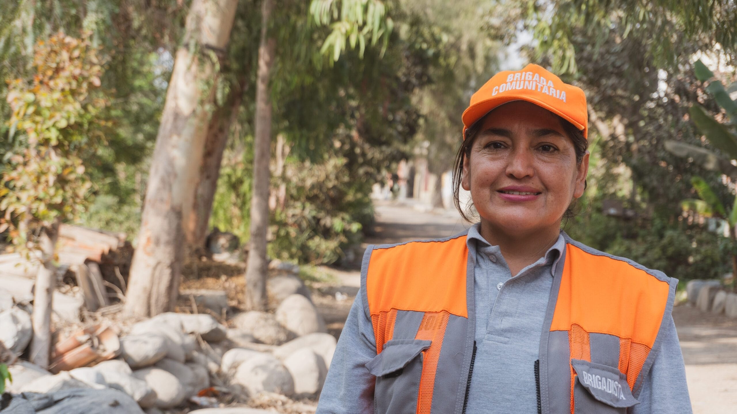 A woman wearing an orange safety vest and cap stands outdoors on a sunny day, smiling. She is in a wooded area with trees, rocks, and bags of rubbish in the background.