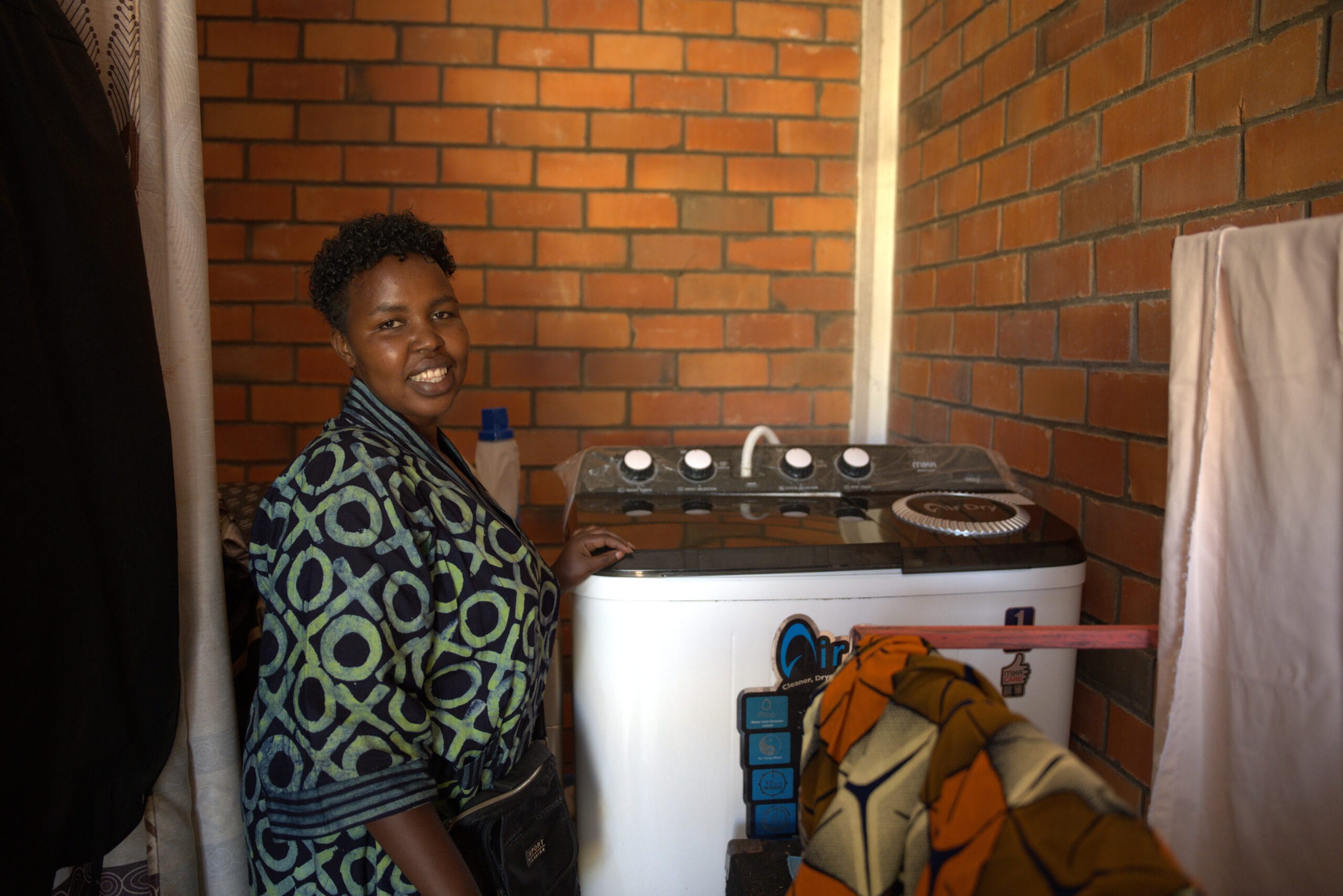 Chance Zawadi - A woman wearing a patterned dressing gown smiles whilst standing next to a top-loading washing machine in a room with brick walls and colourful fabrics hanging nearby.